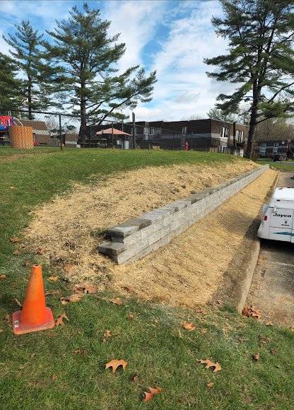 A grassy outdoor area with a newly built stacked stone retaining wall. There is an orange traffic cone on the grass, and a white truck is parked nearby. In the background, there are trees and residential buildings.