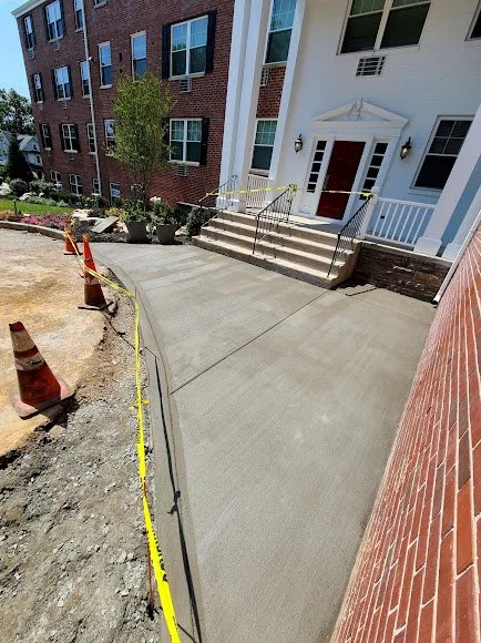 Newly poured concrete pathway leading to the front entrance of a white residential building, with orange construction cones and caution tape on the side under construction.