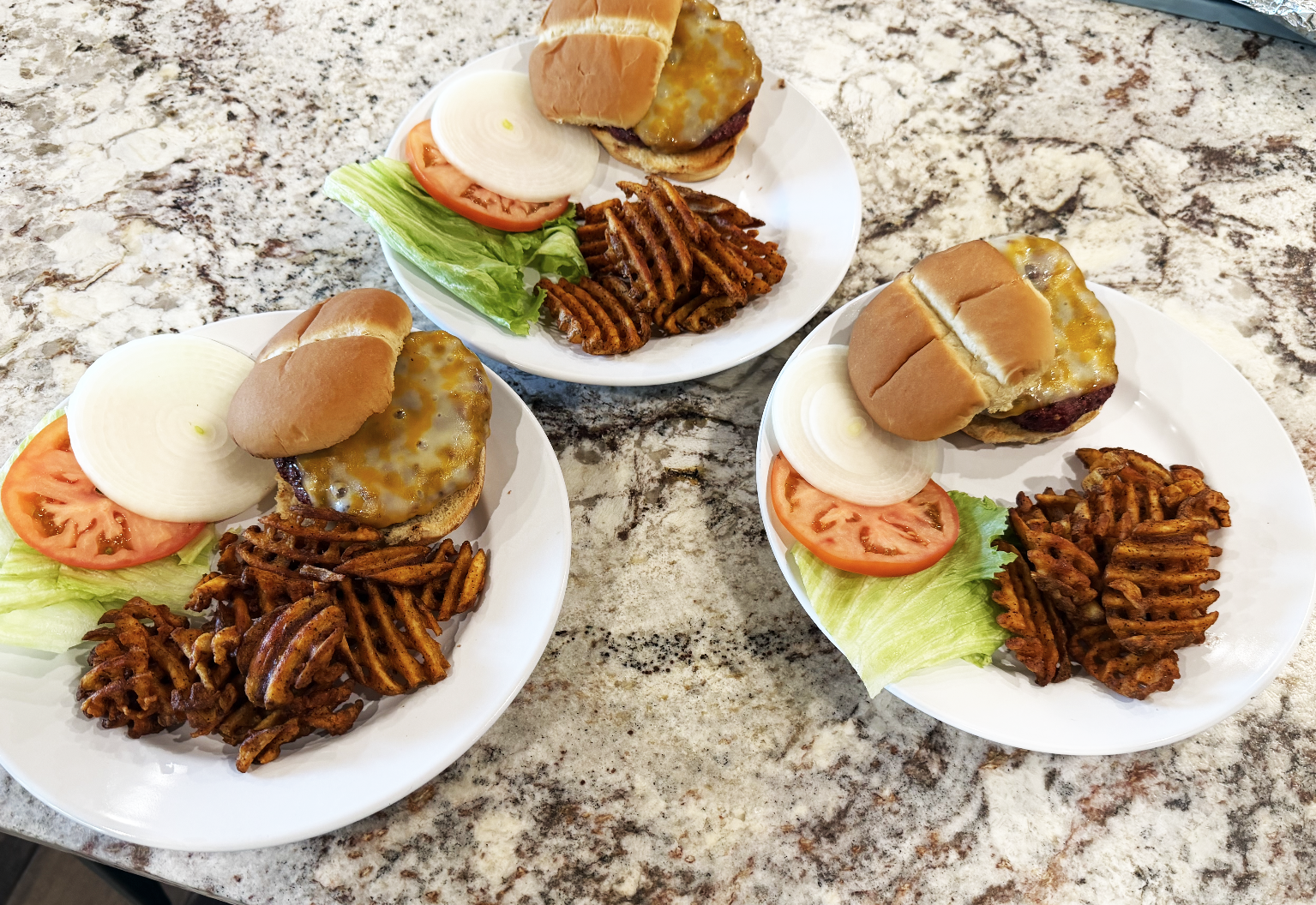 Three plates of Addax sliders with lettuce, tomato, onion, waffle fries, on a granite countertop.