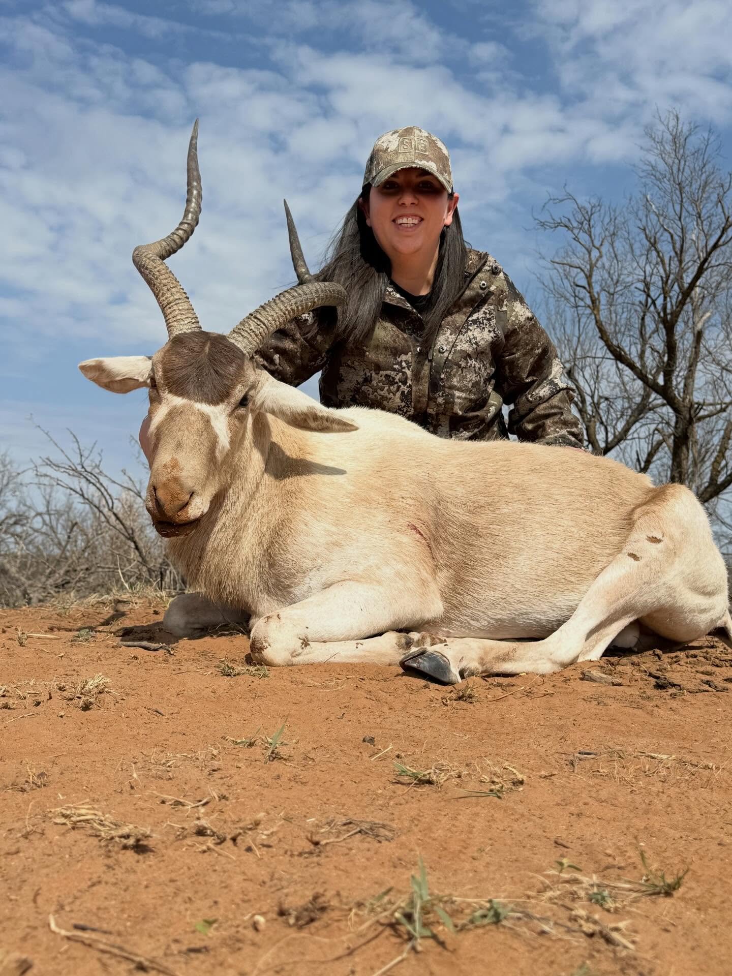 A woman in camouflage clothing kneels next to a Addax with curved horns on bare ground, with a background of leafless trees and a partly cloudy sky.