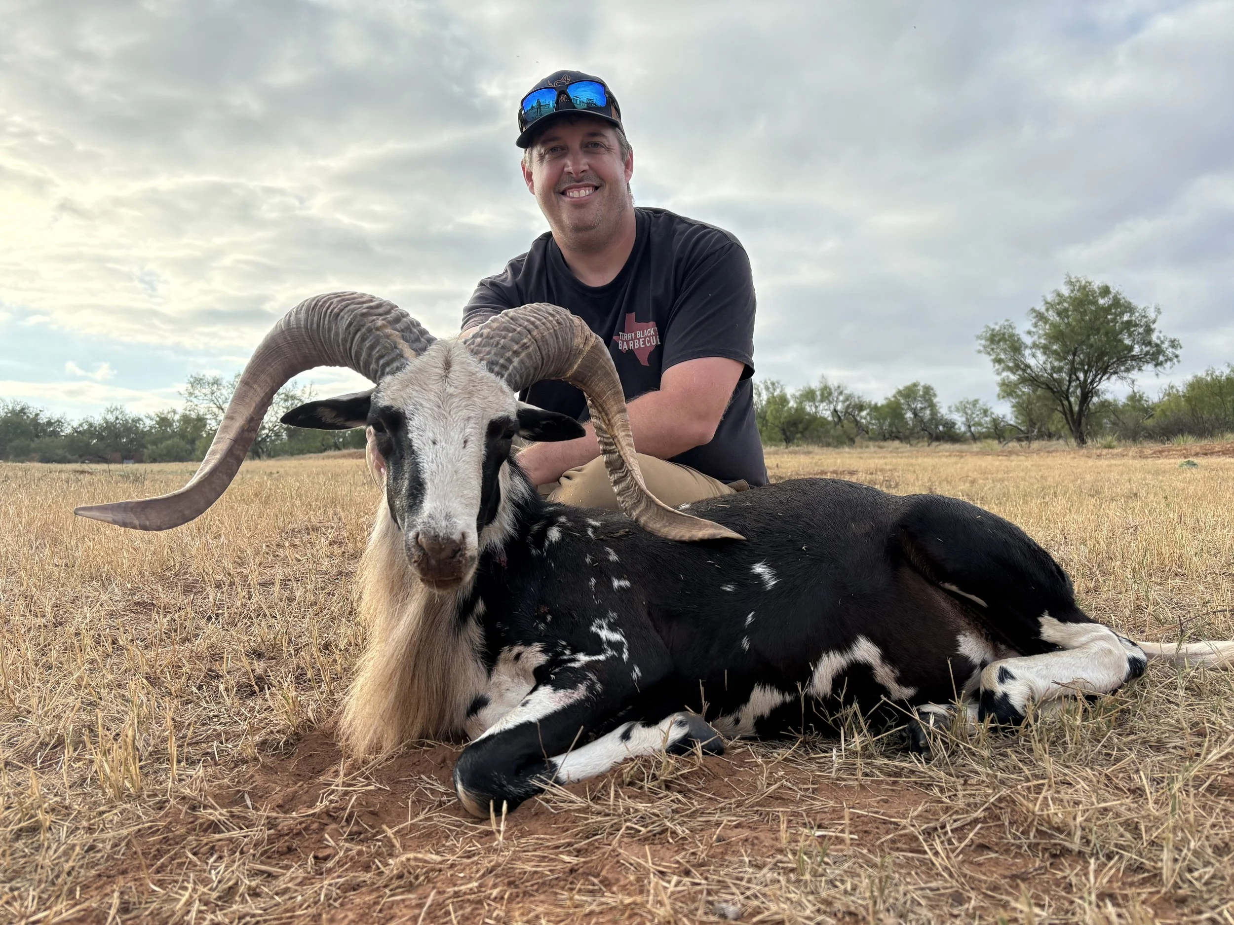 A man smiling and wearing sunglasses and a black T-shirt with a Texas map on it, is kneeling on the ground in an open field with a black and white goat with large, curved horns in front of him and hanging onto his arm. The sky is partly cloudy, and there are trees in the background.
