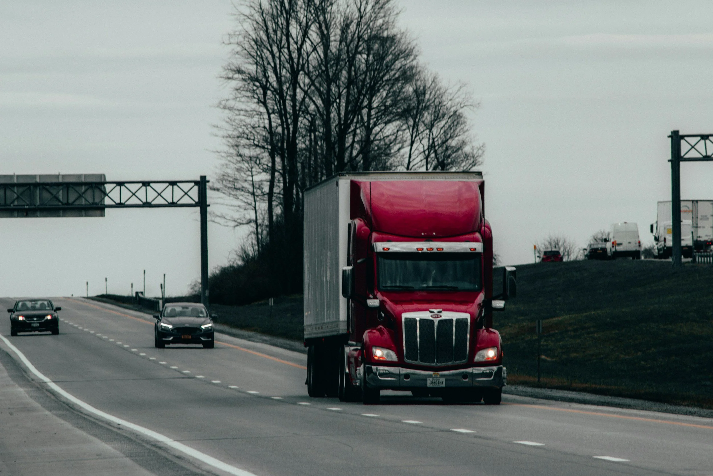 A red semi-truck driving on a highway with other cars and trucks in the background, under an overcast sky with leafless trees.