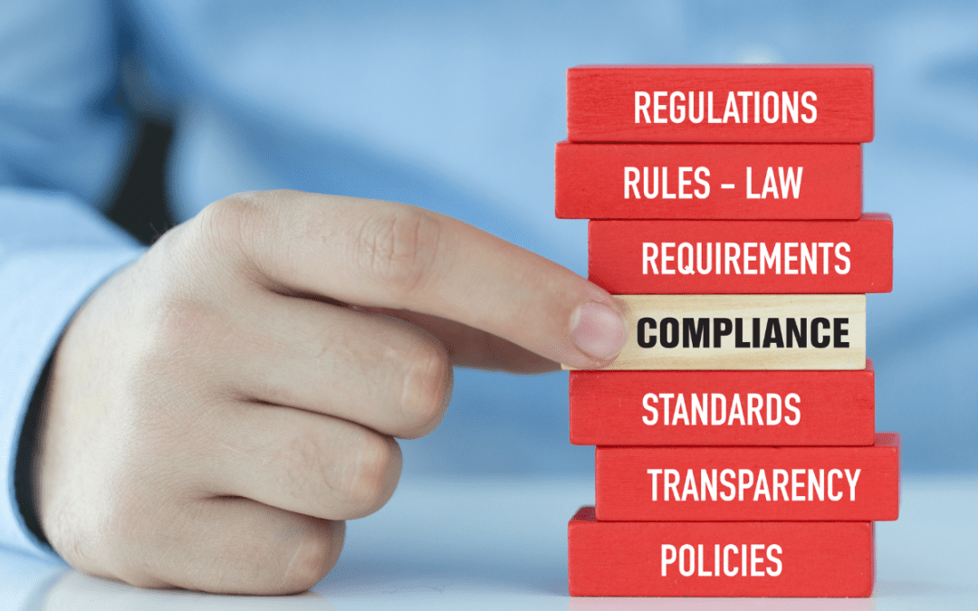 A hand pointing to a stack of red and beige blocks labeled with terms such as 'Regulations,' 'Rules — Law,' 'Requirements,' 'Compliance,' 'Standards,' 'Transparency,' and 'Policies,' with the word 'Compliance' highlighted in black.
