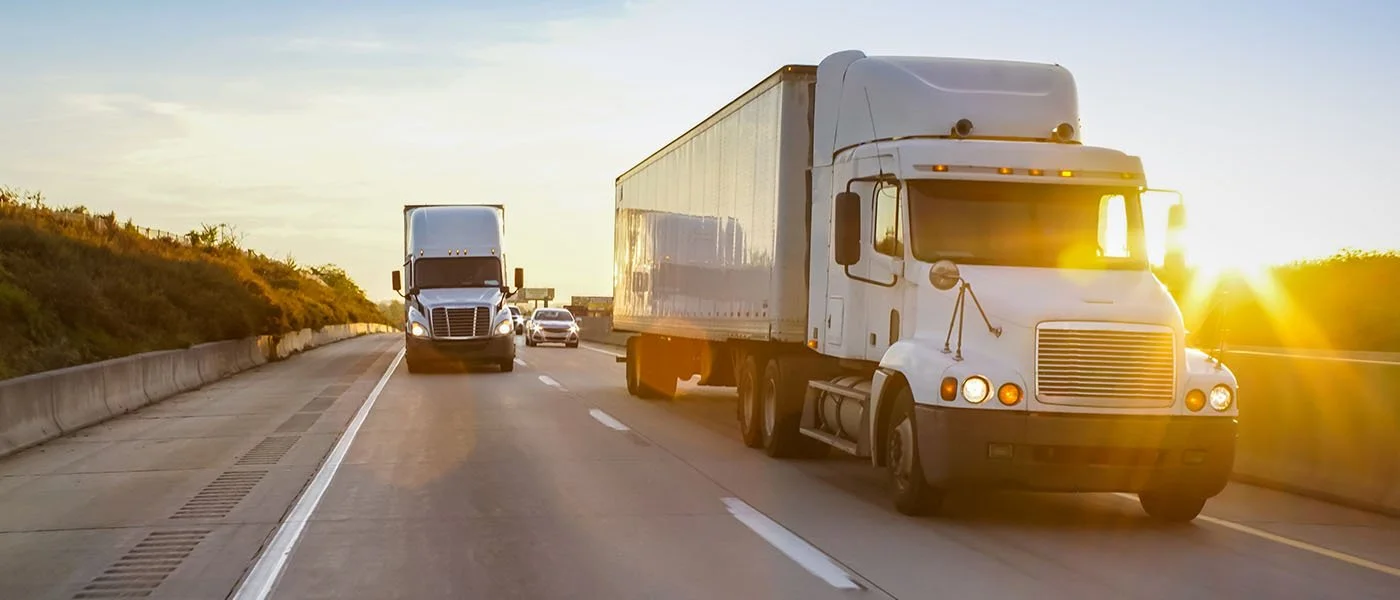 White semi-truck driving on highway during sunset with other vehicles, including a black truck and cars, on a scenic road with greenery on the side.