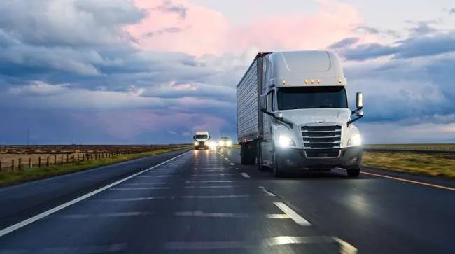 A semi-truck driving on a wet highway during dusk with other vehicles visible in the distance.