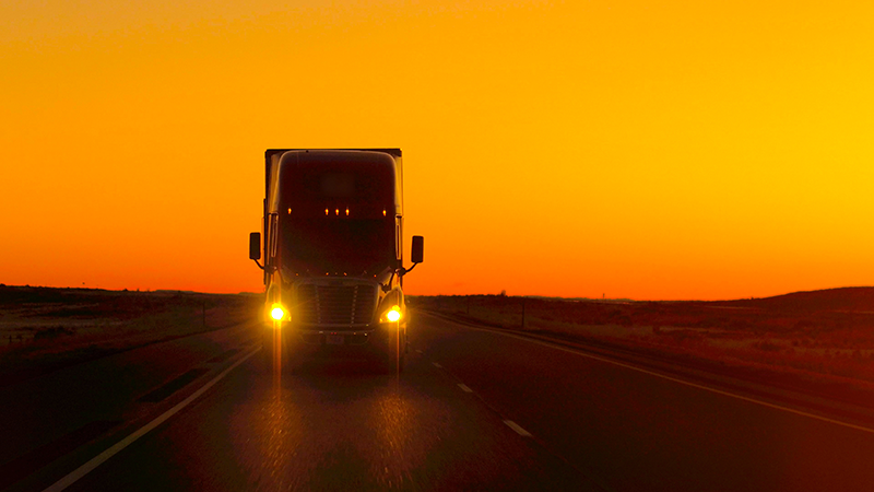 A large truck driving on an empty road at sunset, with the sun setting behind it and its headlights on.