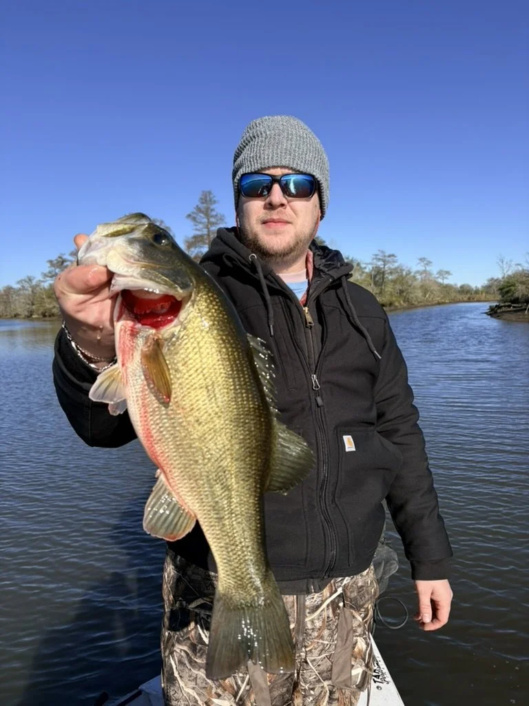 Man in a beanie, sunglasses, and black jacket holding a large bass fish near a lake on a clear day.