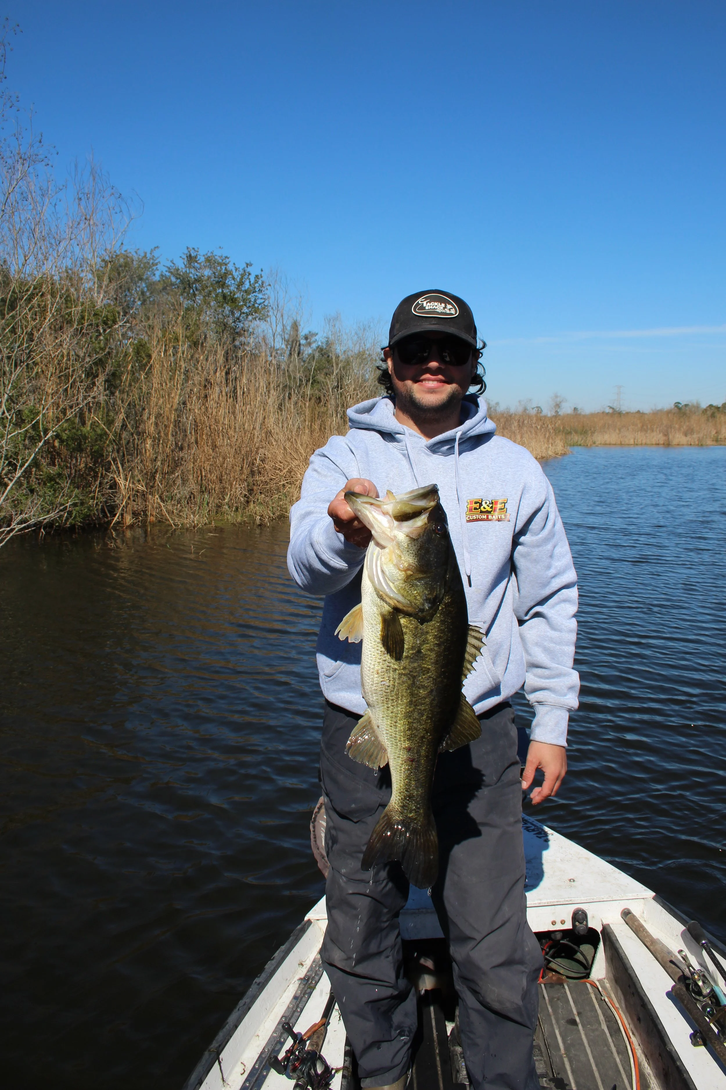 A man standing in a boat holding a large fish, wearing a gray hoodie, black cap, sunglasses, and dark pants, on a river with tall grass and trees in the background under a clear blue sky.