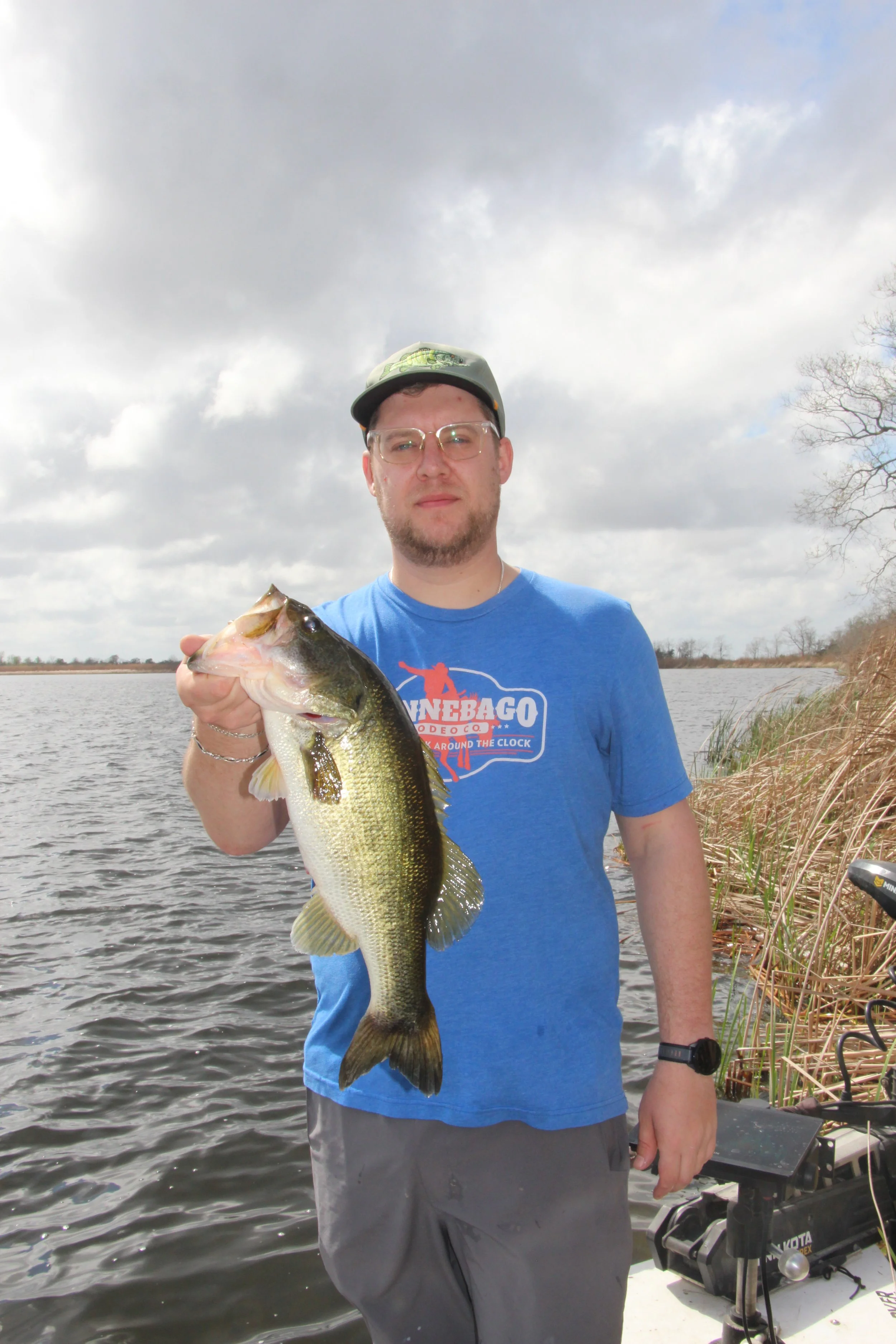 A man wearing a baseball cap, glasses, and a blue T-shirt holding a large fish that he caught in a body of water, with cloudy sky and some trees in the background.