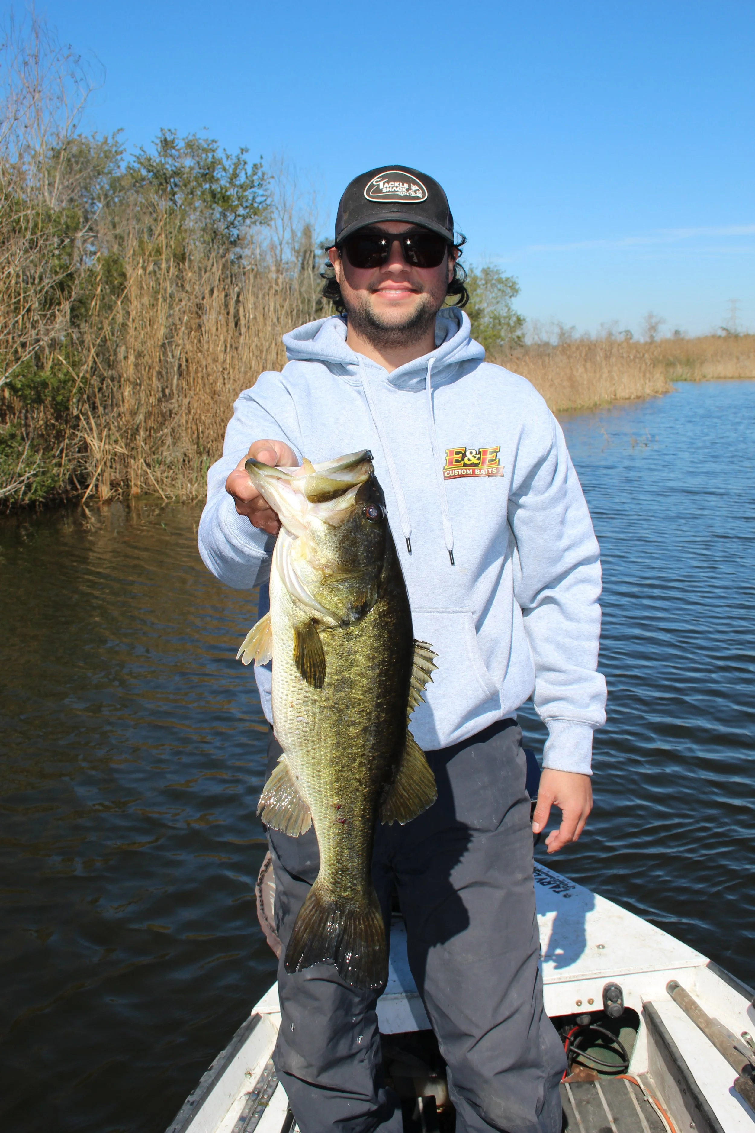 A man in sunglasses and a hoodie holding a large bass fish on a boat in a river with grassy banks and a clear blue sky.