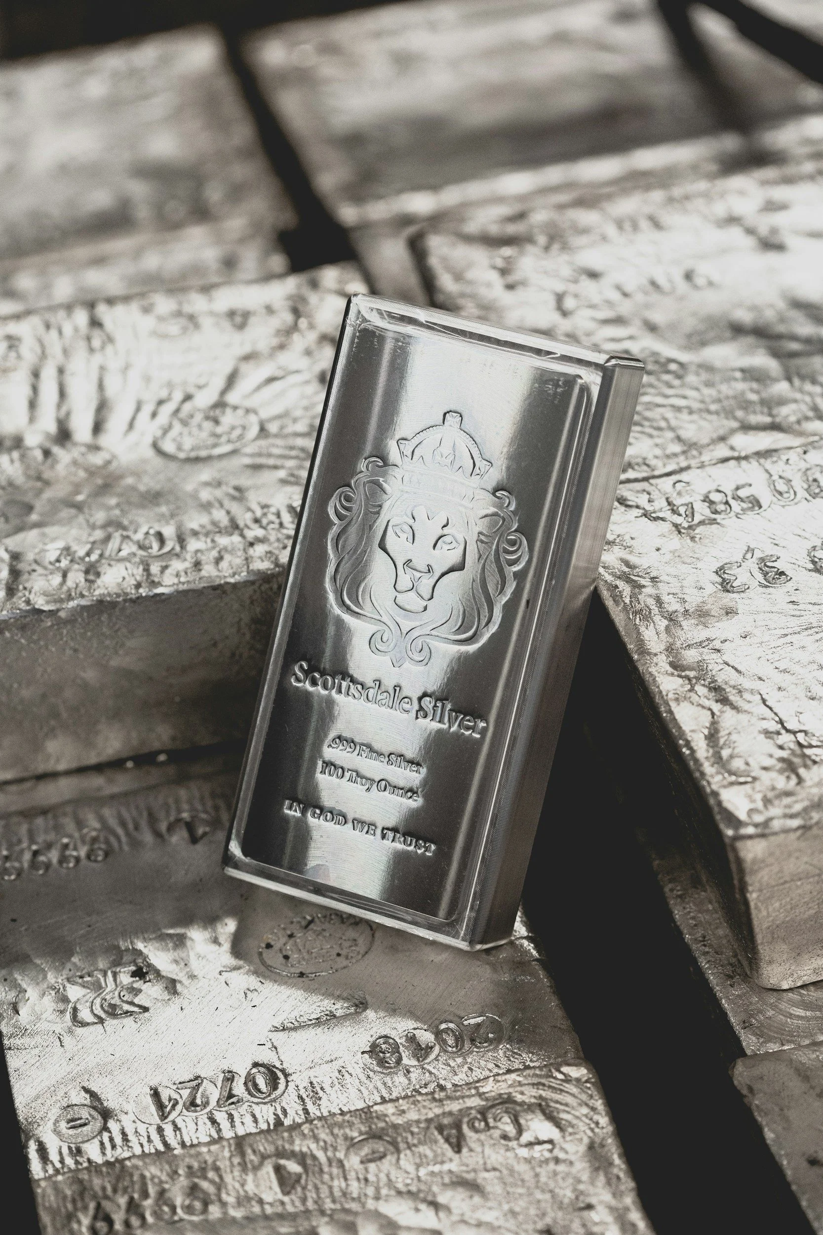 A silver bar with a lion's head engraving and the inscription "Scottsdale Silver, 999 Fine Silver, 100 Troy Ounces, In God We Trust" on its surface, leaning against other silver bars.