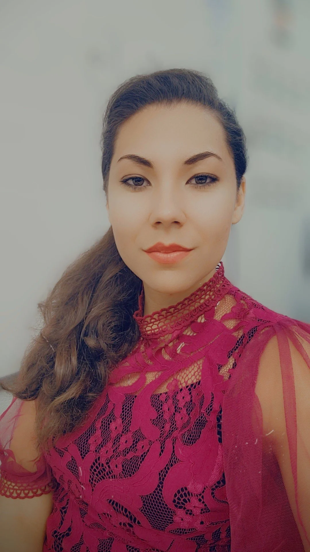 A woman with long, wavy hair wears a red, lace top with intricate floral patterns and a high, lace neckline.