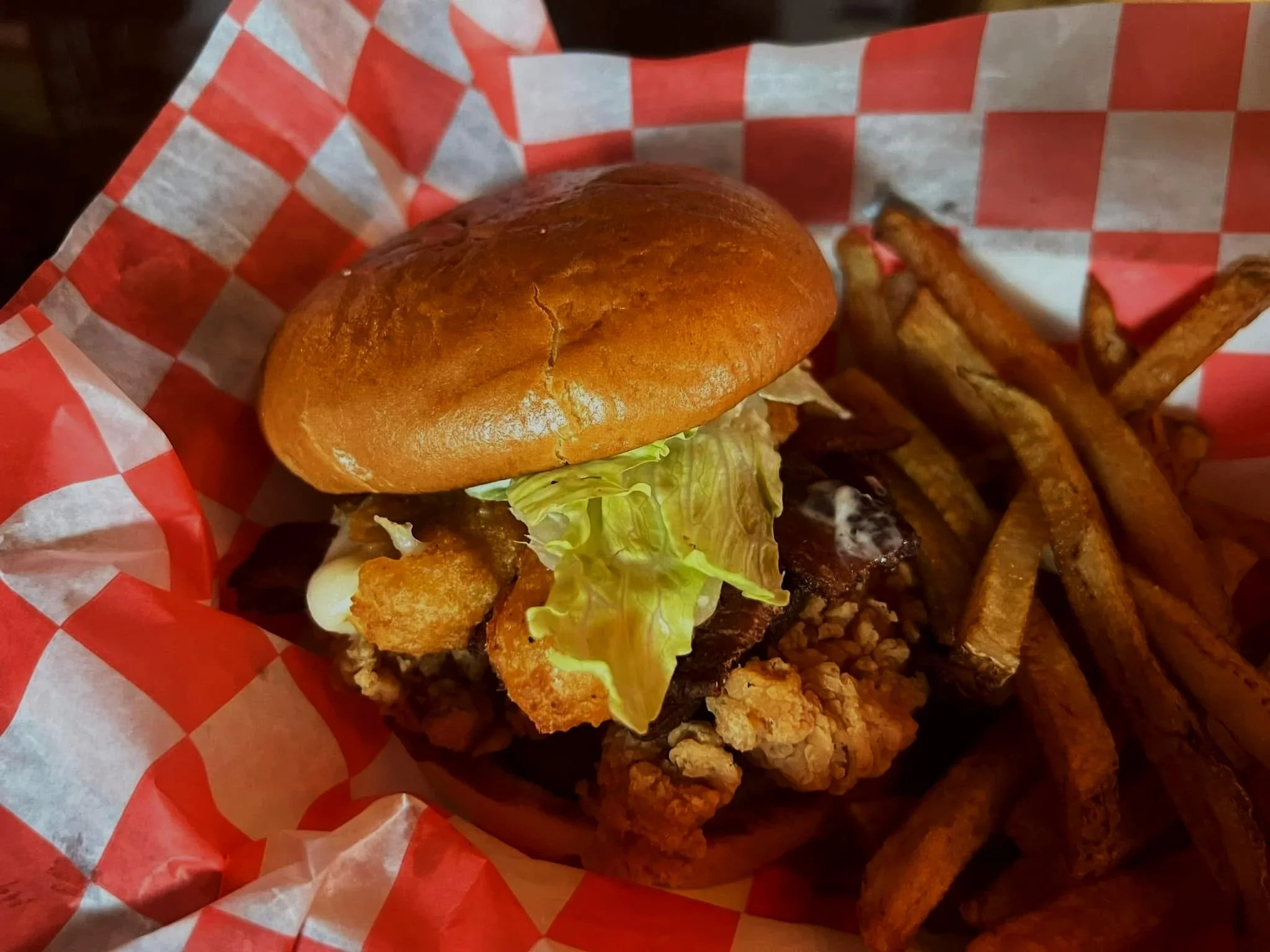 Fried chicken sandwich with lettuce, on a toasted bun, served with French fries on red and white checkered paper.