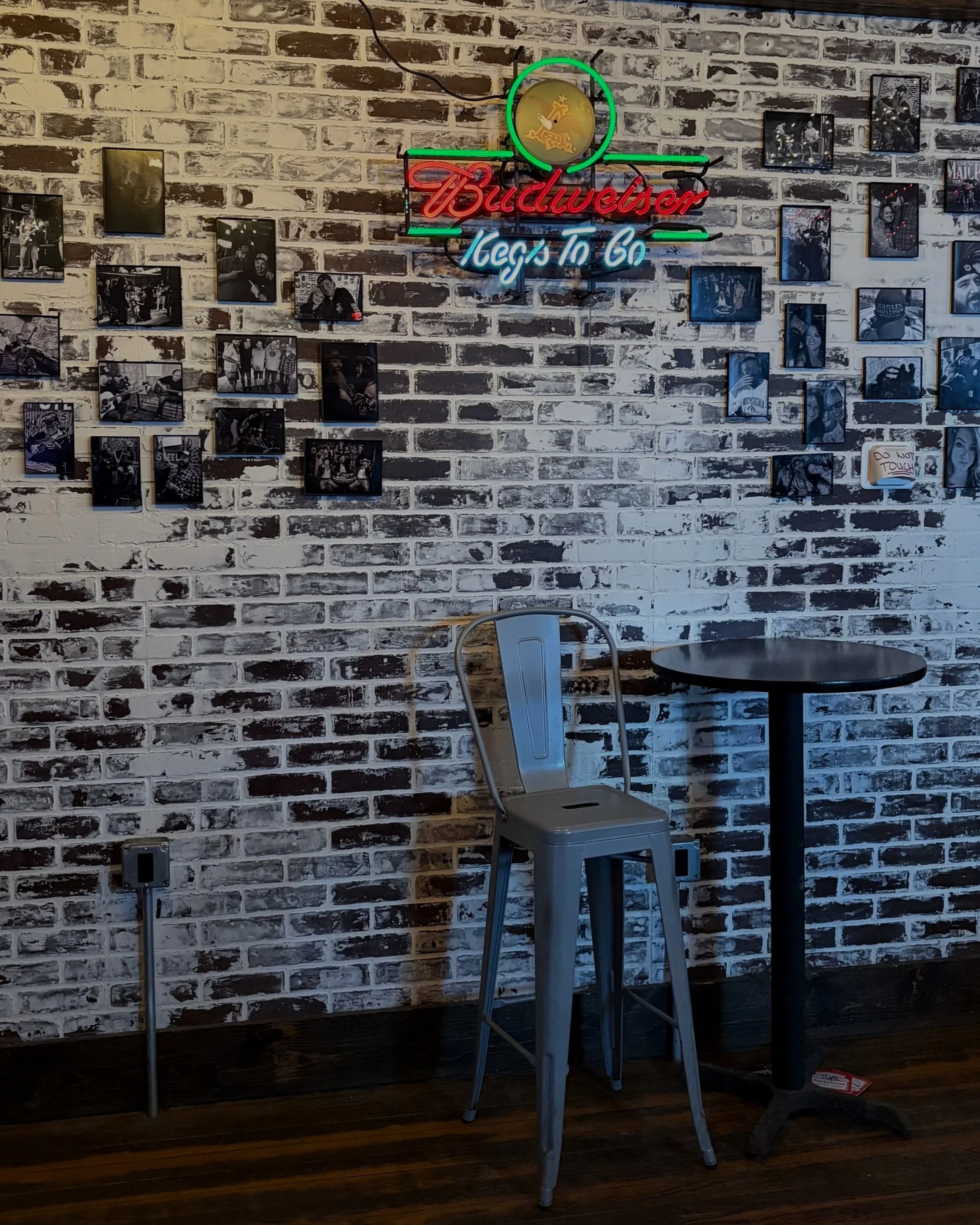 Interior of a bar with a brick wall decorated with black-and-white photos of great memories at Kenny B's, a neon sign that says 'Budweiser Let's To Go,' a silver chair, and a black round table.