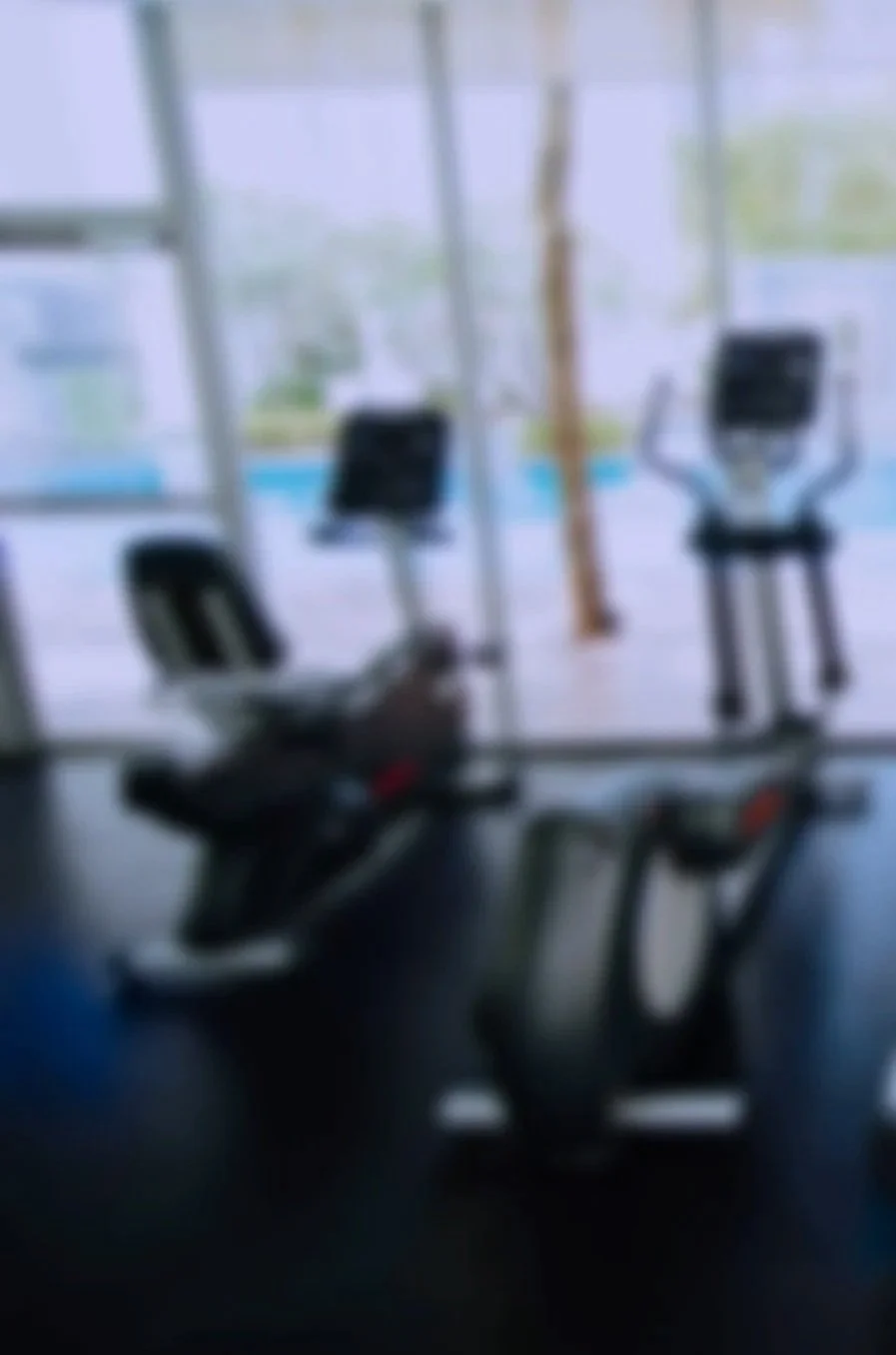 Indoor gym with two exercise machines, a recumbent bike and an elliptical trainer, near large glass windows that overlook a pool and garden outside.