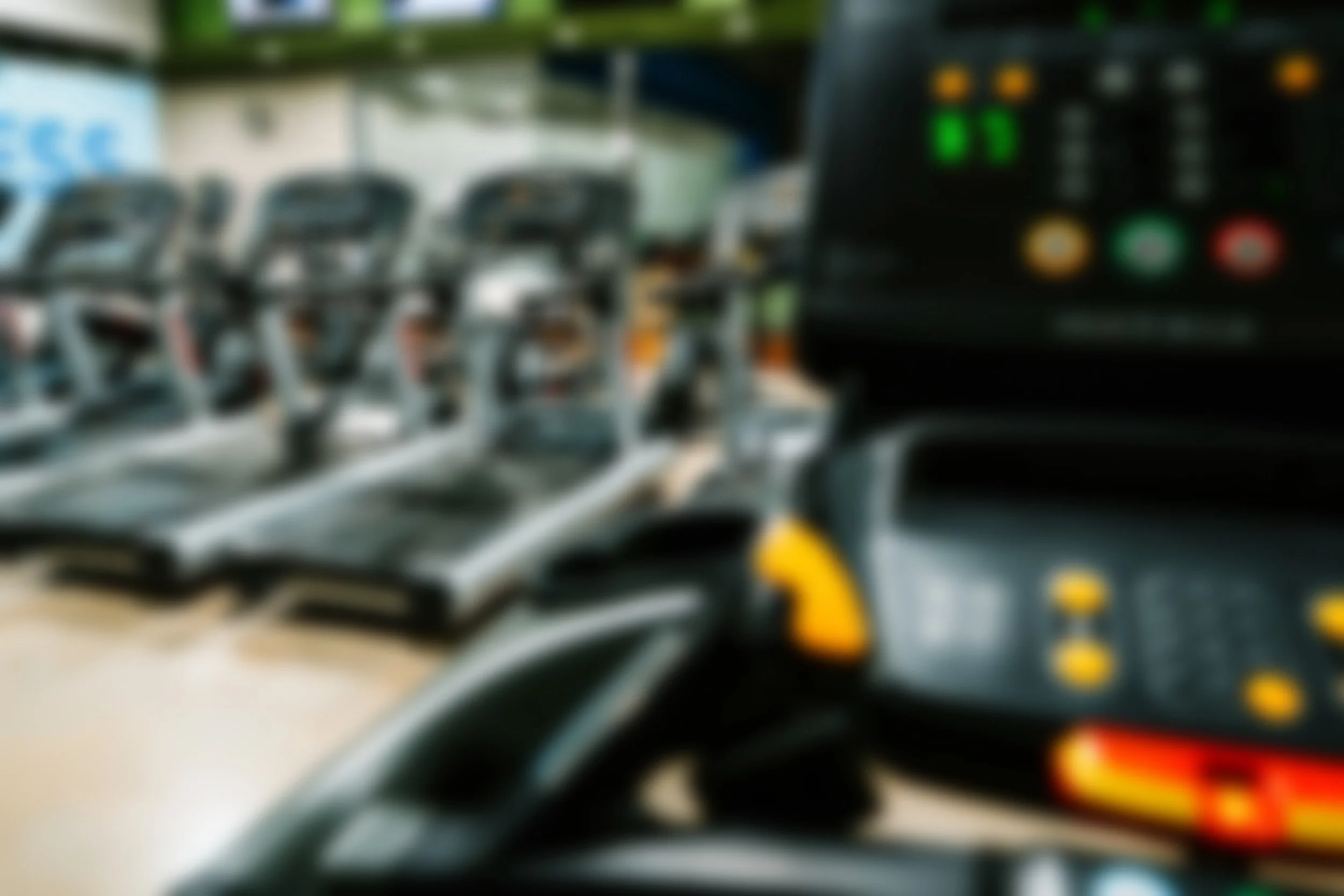 An indoor gym with multiple treadmills in a row, a mirror wall, a clock, and screens displaying information. A fitness tracker or remote control is partially visible in the foreground.