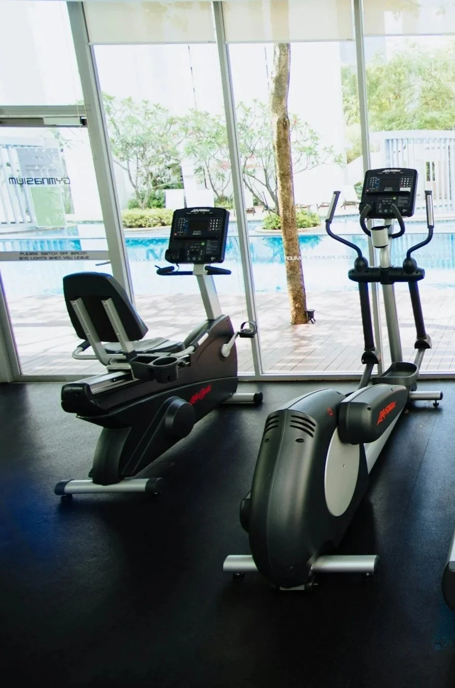 Indoor gym with two exercise machines, a recumbent bike and an elliptical trainer, near large glass windows that overlook a pool and garden outside.