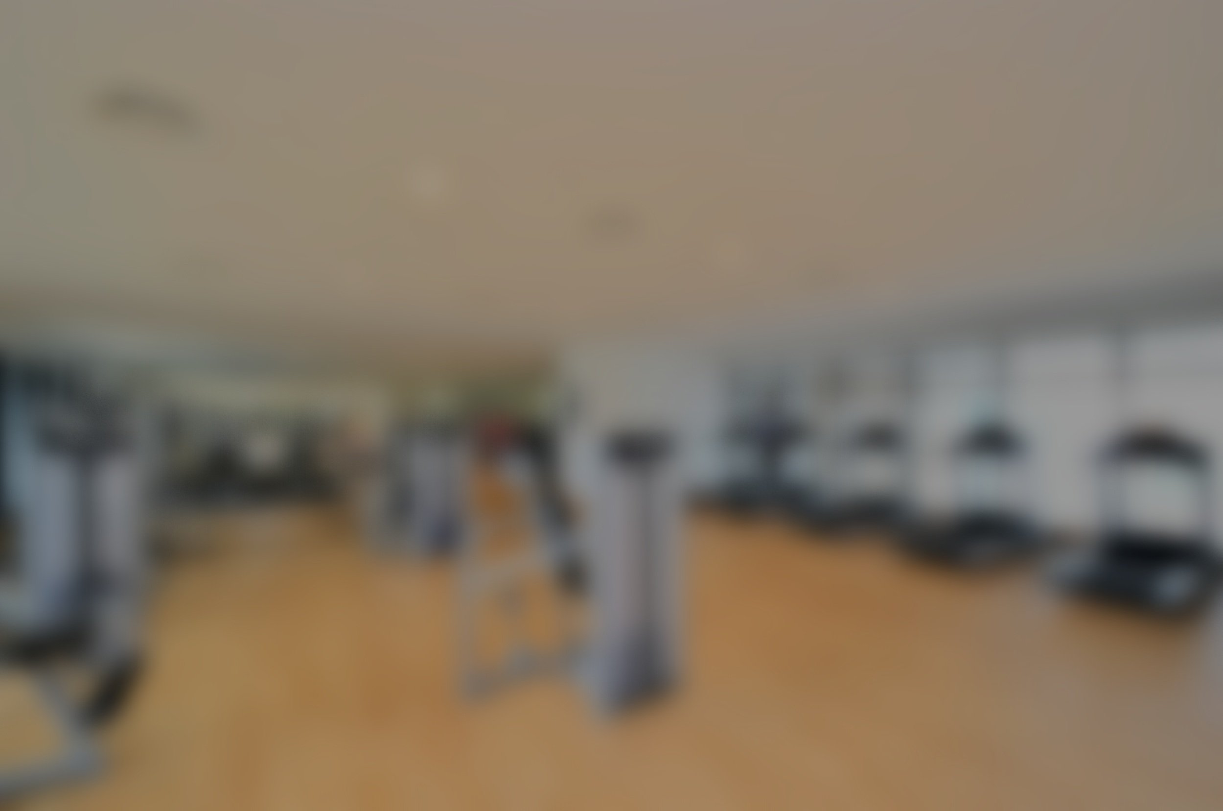 Empty gym with four treadmills in the foreground, weightlifting equipment, dumbbells, exercise bikes, and a bench in the background, and large windows with natural light.