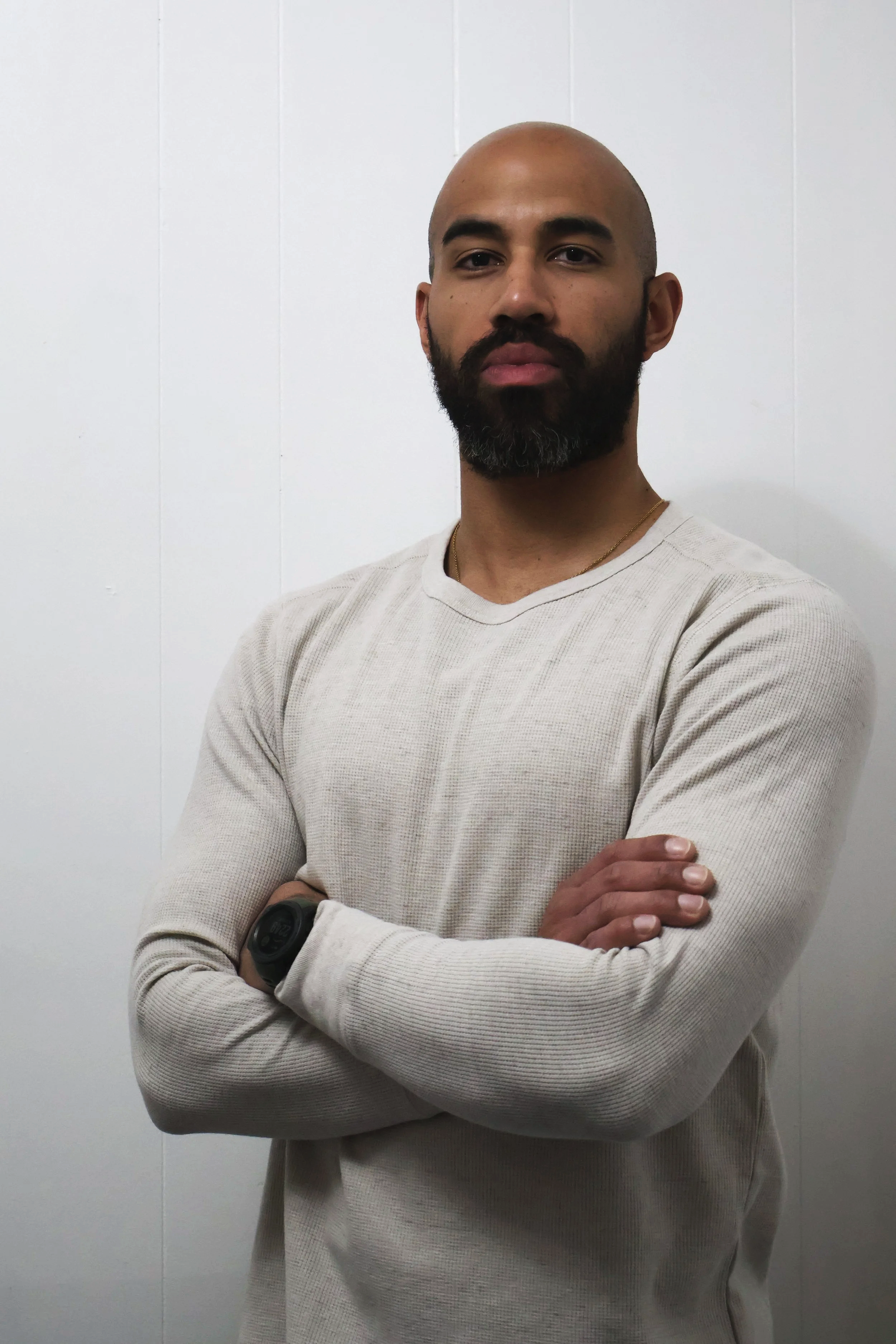 A man with a beard and crossed arms, wearing a beige long-sleeve shirt and a watch, standing against a plain white wall.