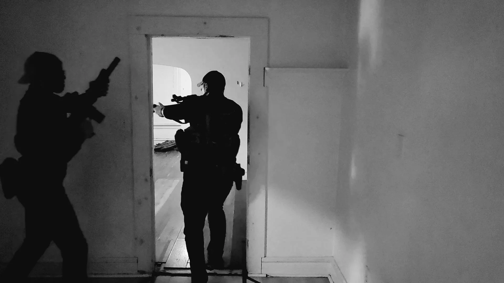 Silhouettes of two armed law enforcement officers during a tactical operation, inside a building, with one pointing a weapon and the other aiming a firearm through a door.