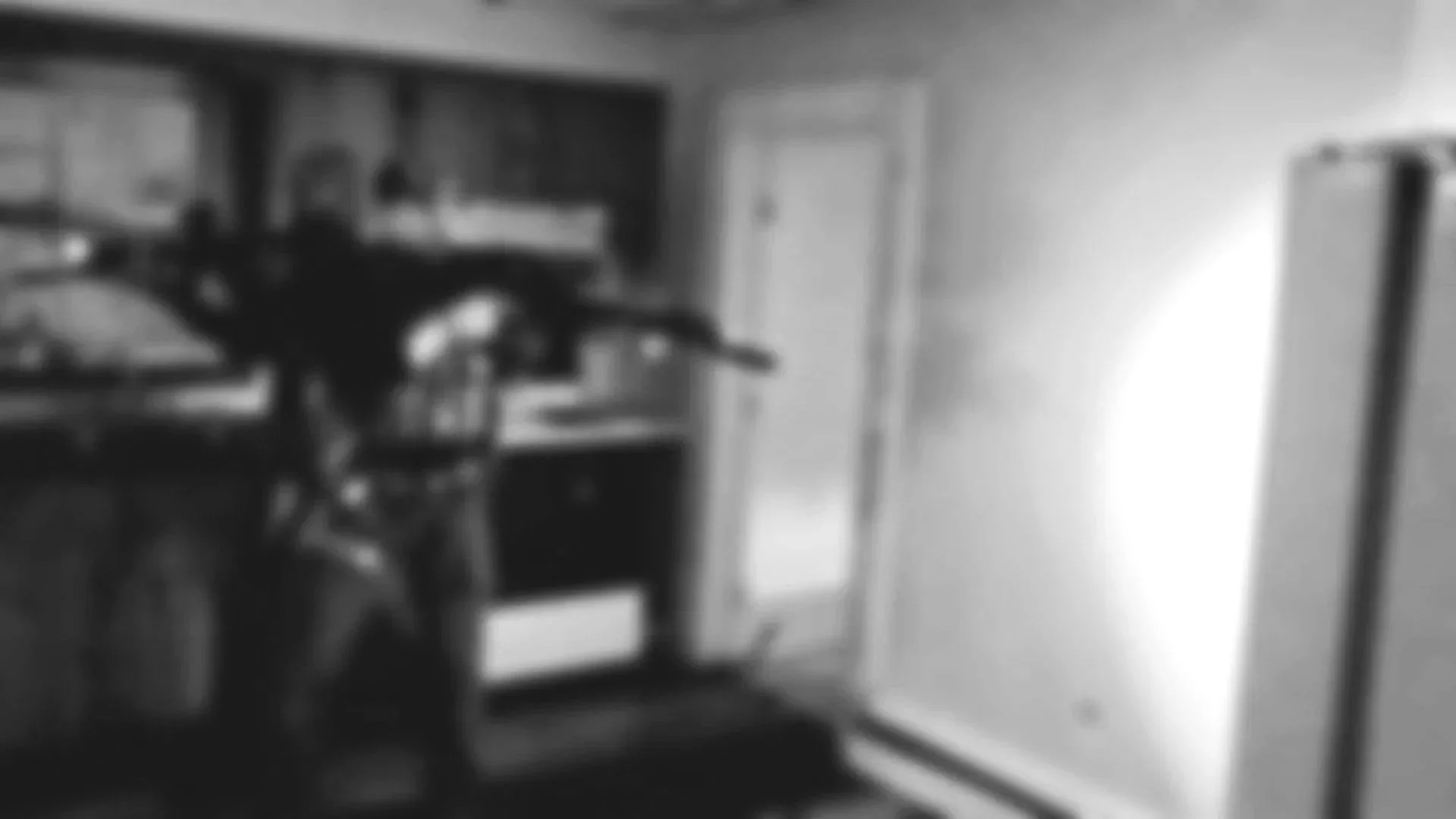 Blurry, black and white image of a room with a door, a desk with a chair, and shelves with objects, possibly a bedroom or office.