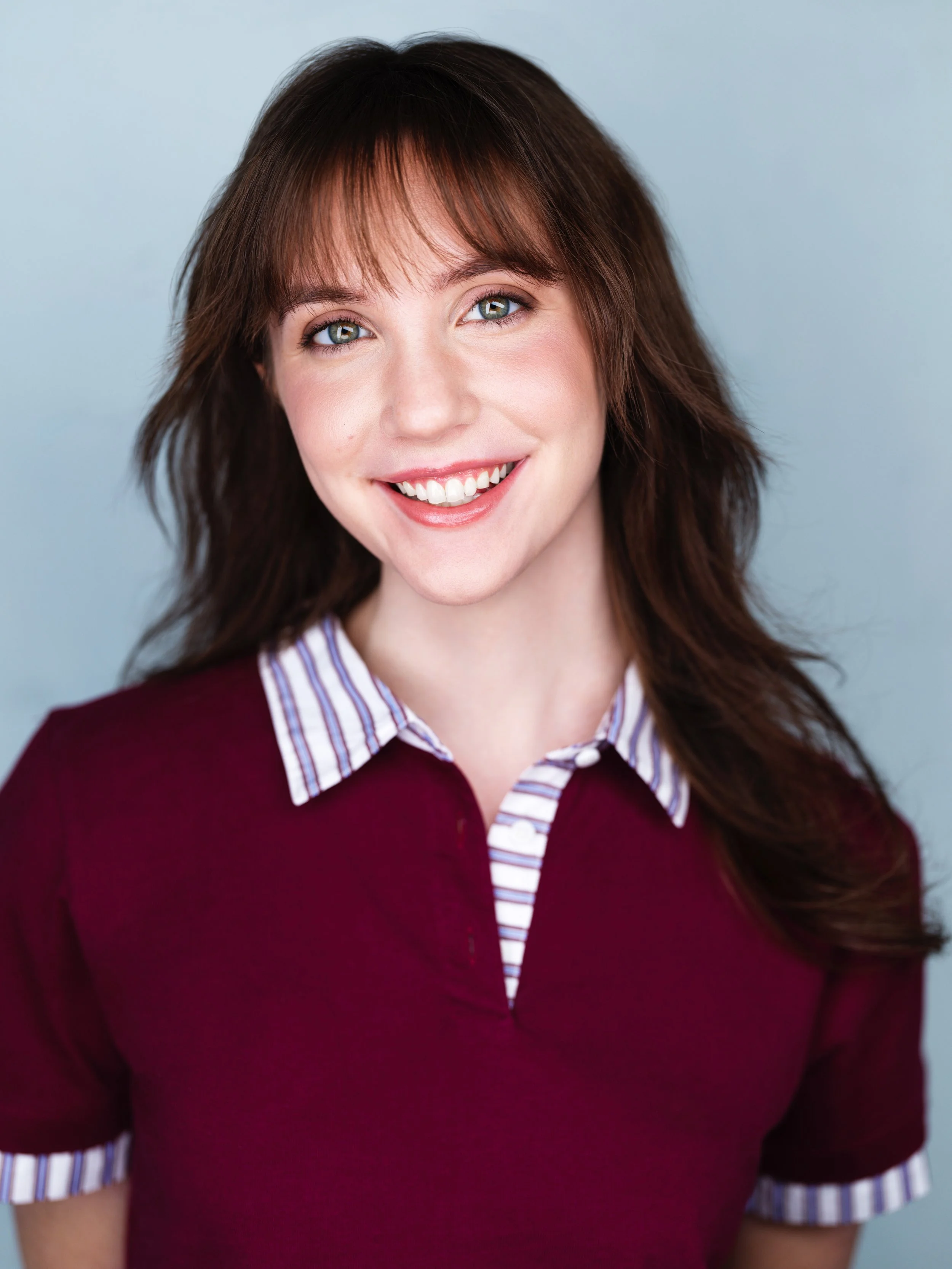 Co-star headshot of a young, GenZ, college-aged actor with light skin, green eyes, and brown hair with bangs, smiling. She portrays a profession-based role.