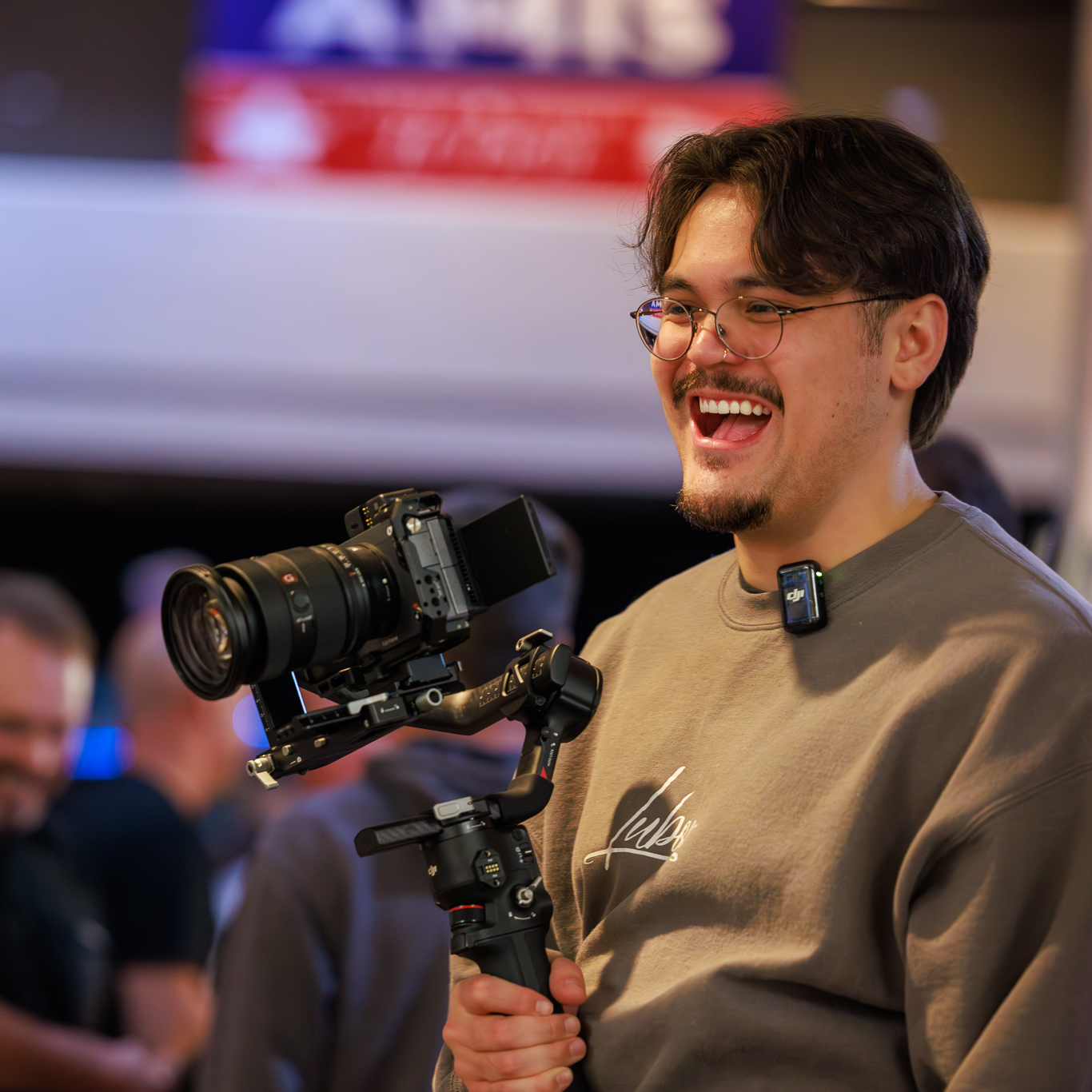 A young man with glasses and a beard holding a professional camera with a stabilizer, smiling and looking to the right at an indoor event.