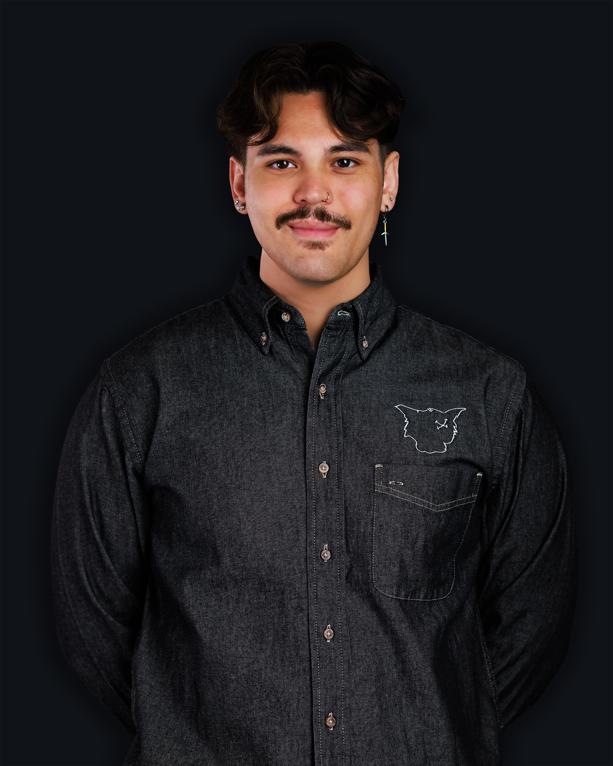 A young man with dark hair, a mustache, and earrings wearing a black button-up shirt with a cat face embroidered on the chest, standing against a black background.
