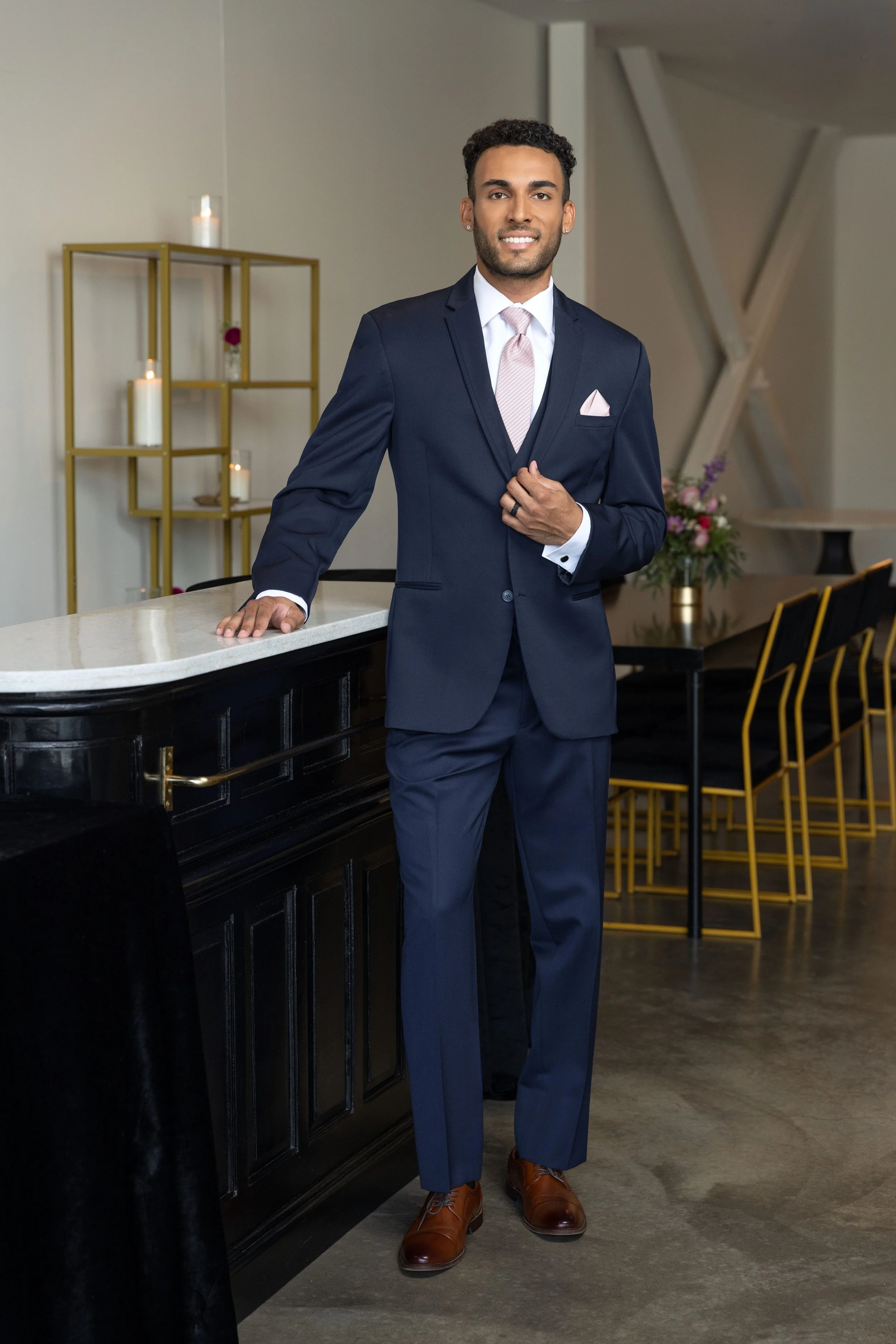 A young man in a navy suit, white shirt, and light pink tie, standing at a bar with his left hand on the black bar counter. He has dark curly hair, a beard, and is smiling at the camera. The background features a modern interior with candles, flowers, and yellow chairs.