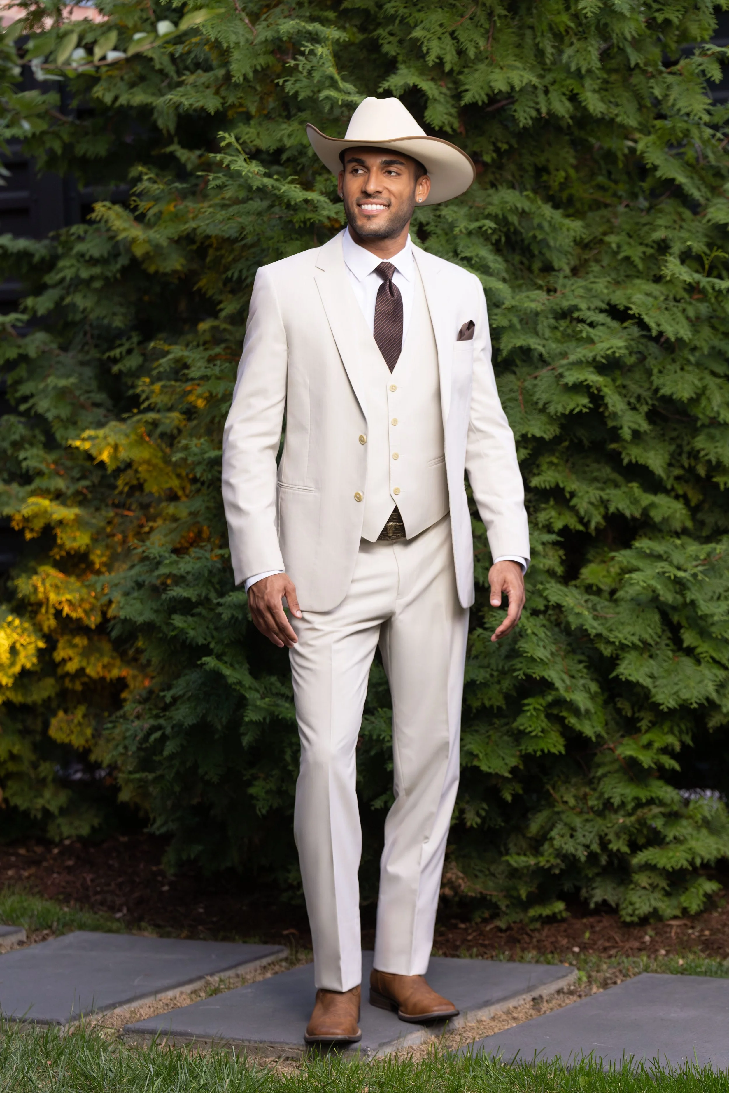 A man standing outdoors on gray stepping stones, wearing a white suit, brown cowboy hat, brown boots, and a patterned tie, with green foliage in the background.