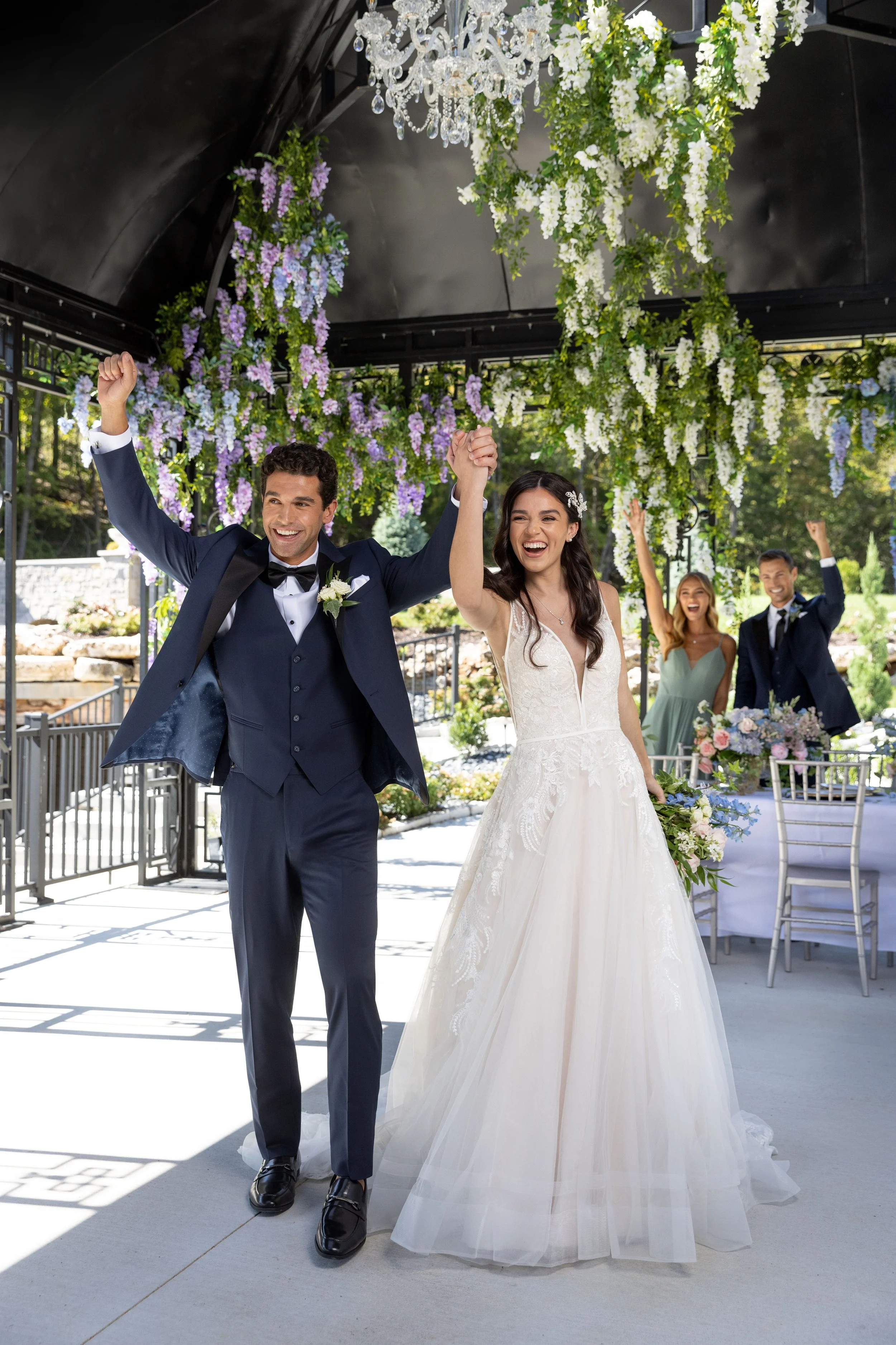 A newlywed couple celebrating at an outdoor wedding ceremony, with the groom in a navy suit and the bride in a white wedding dress, holding hands and smiling with guests in the background, decorated with hanging white and purple flowers and a chandelier.