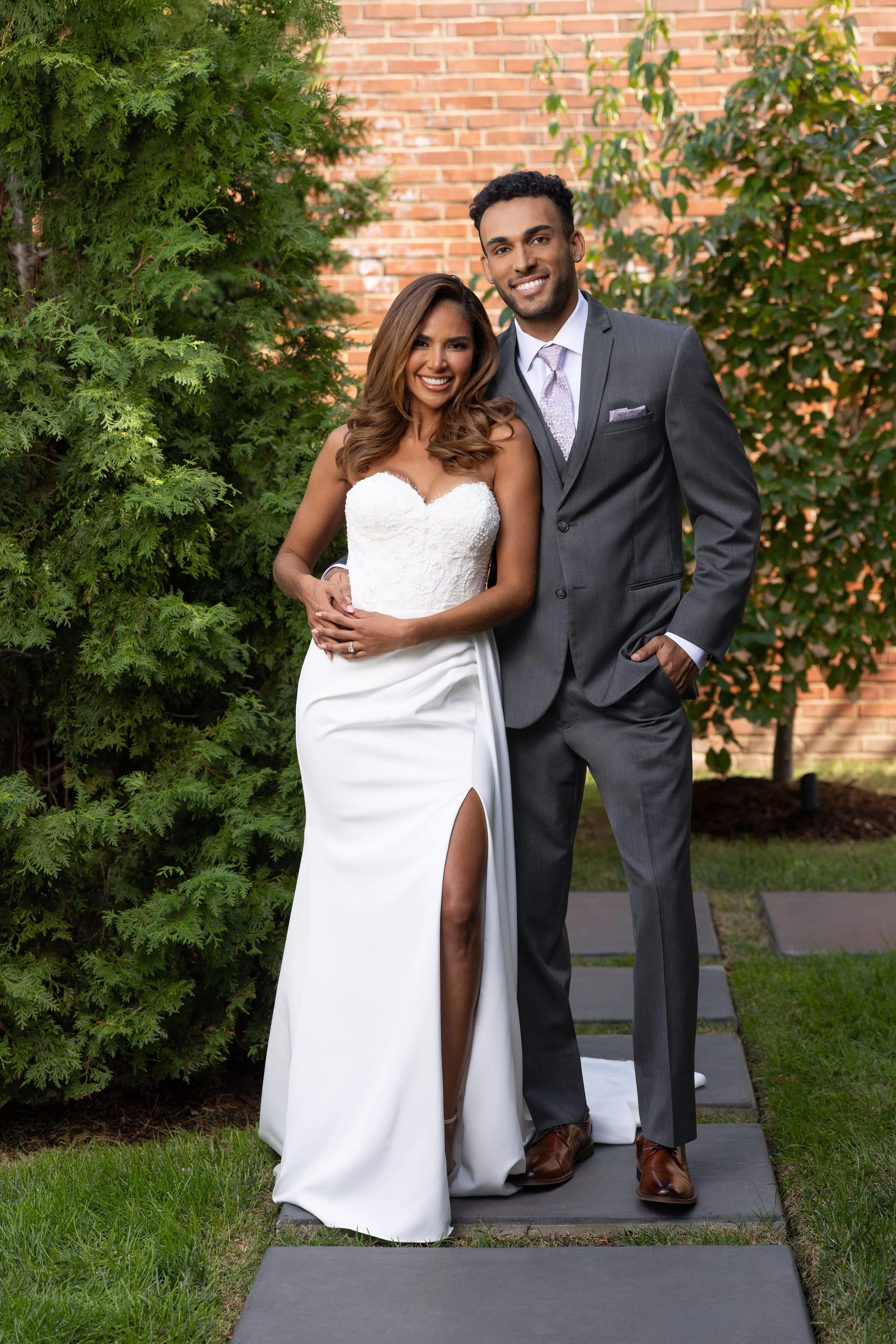 A smiling couple dressed in wedding attire standing outdoors near greenery and a brick wall, with the woman in a white gown and the man in a dark gray suit.