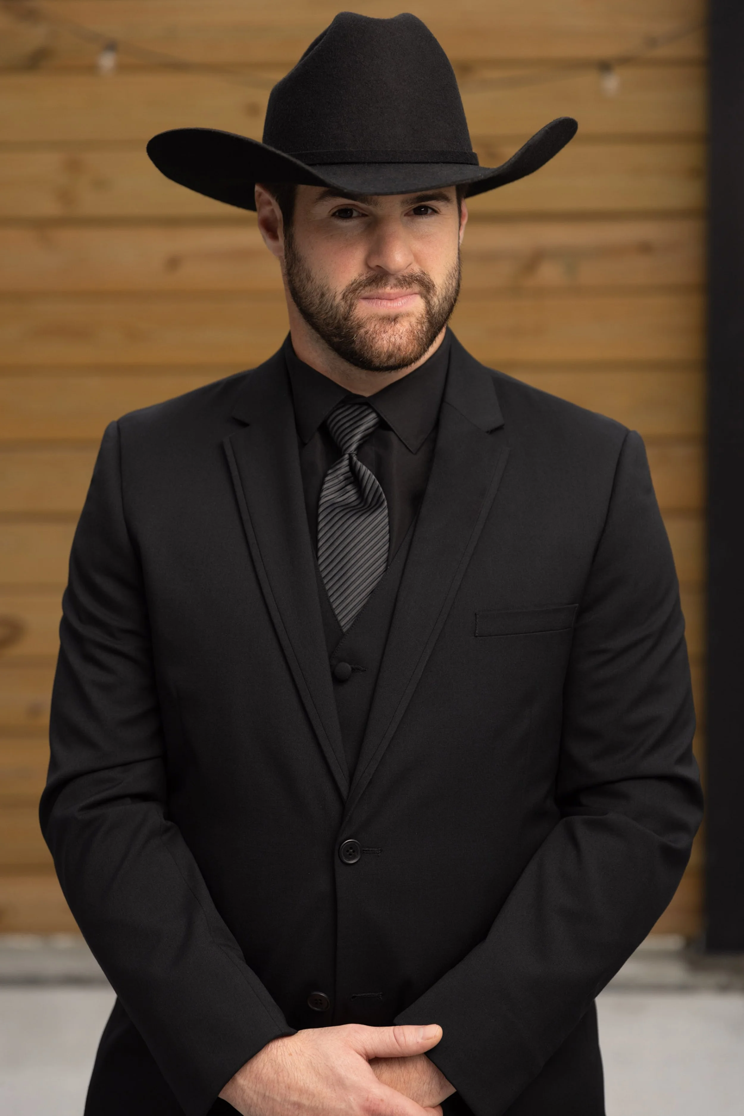 A man wearing a black suit, black tie, and black cowboy hat, standing with arms crossed in front of a wooden wall.