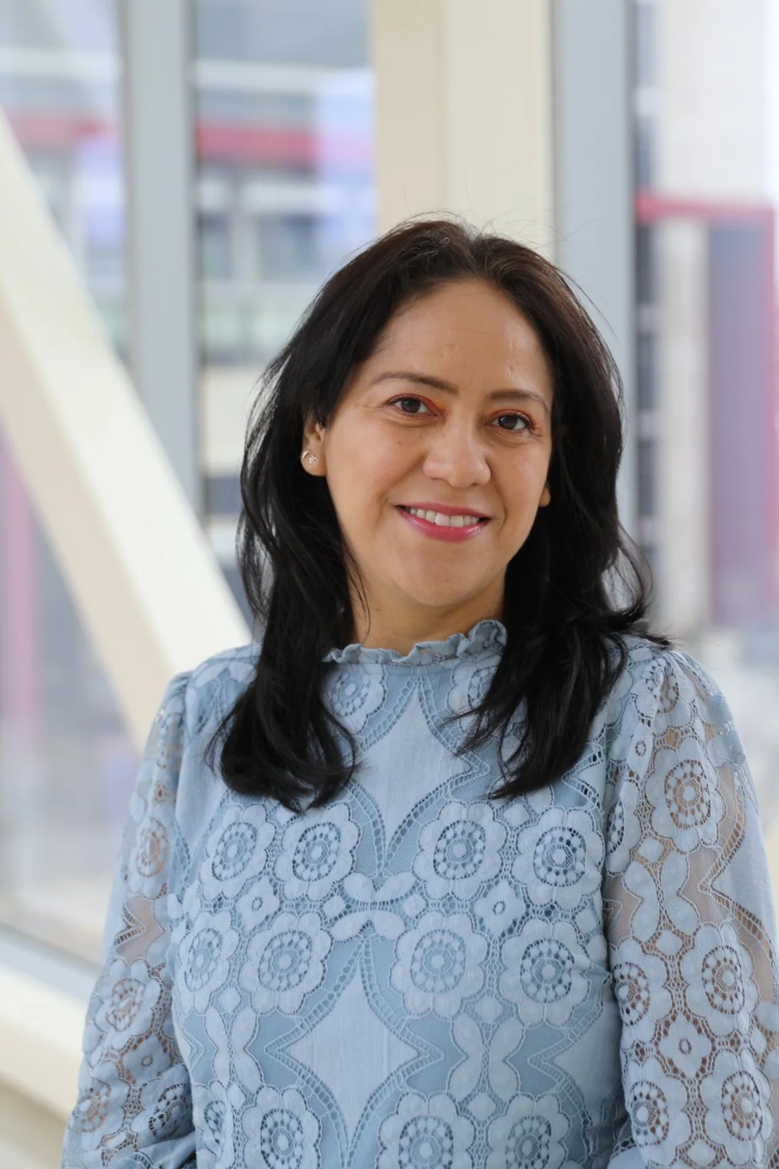 A woman with dark hair smiling, wearing a light blue lace top, standing indoors with windows and buildings in the background.