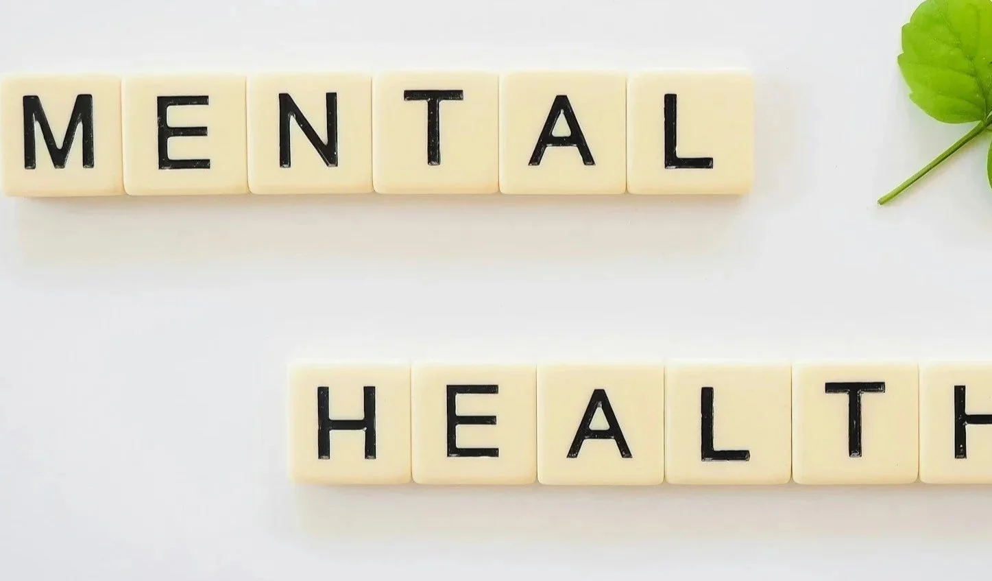 Scrabble tiles spell out 'MENTAL' and 'HEALTH' on a white background, with a green leaf in the top right corner.