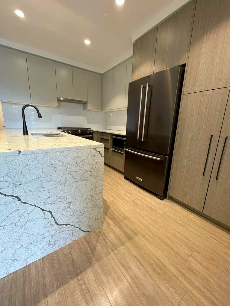 Modern kitchen with beige marble island, black refrigerator, wooden cabinets, and light wooden flooring.