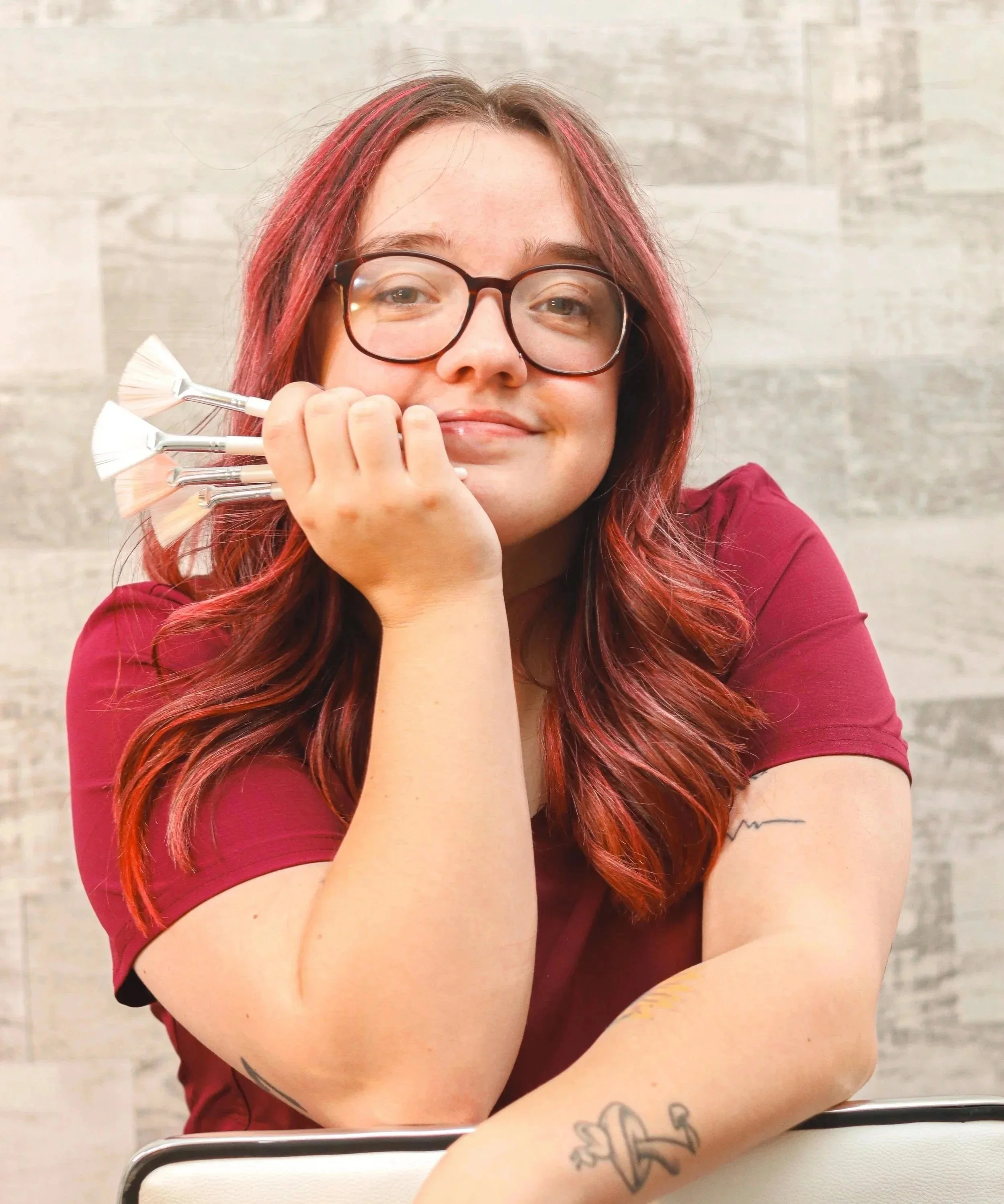 Woman with pink hair and glasses sitting at a table, holding brushes against cheek, smiling, in a room with light wooden wall.