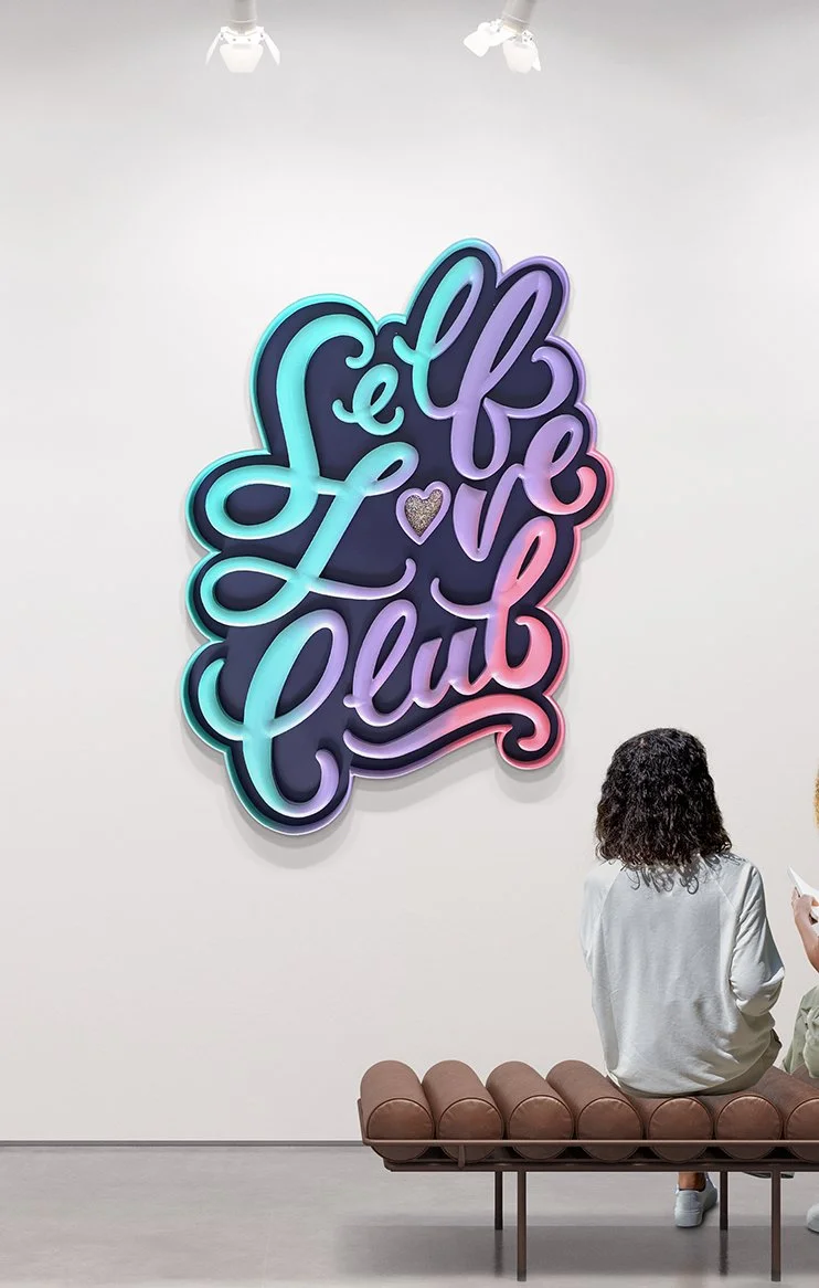 People sitting on a brown bench in front of a colorful sign that reads 'Self Love Club' with a heart in the center, in a gallery or exhibition space.