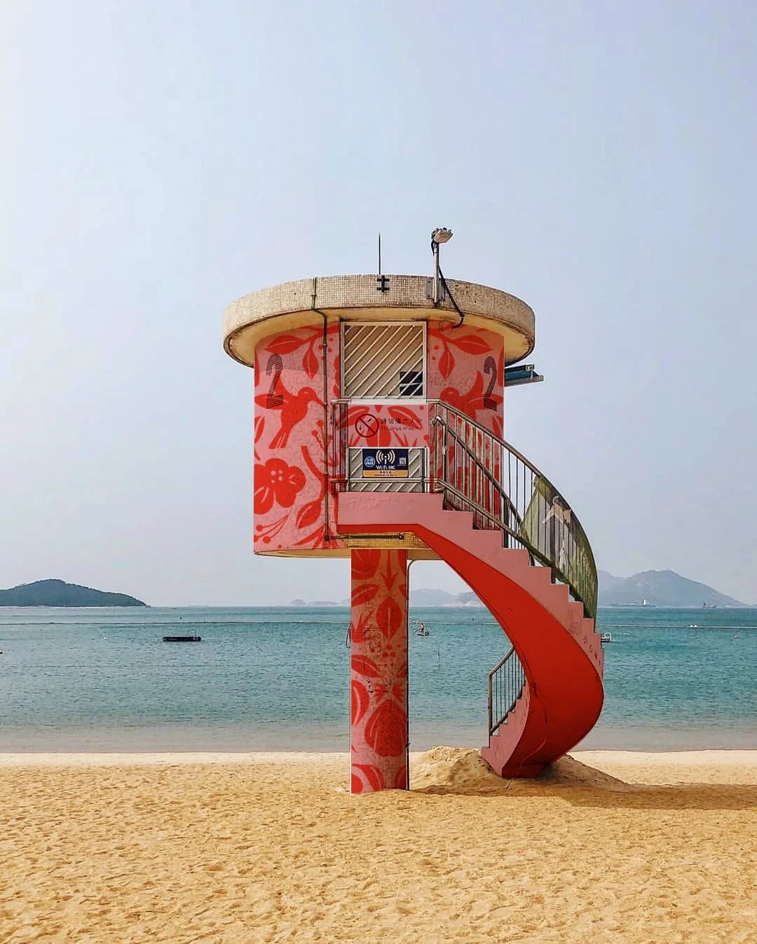A colorful lifeguard tower in Hong Kong with a spiral staircase on a sandy beach with water and islands in the background.