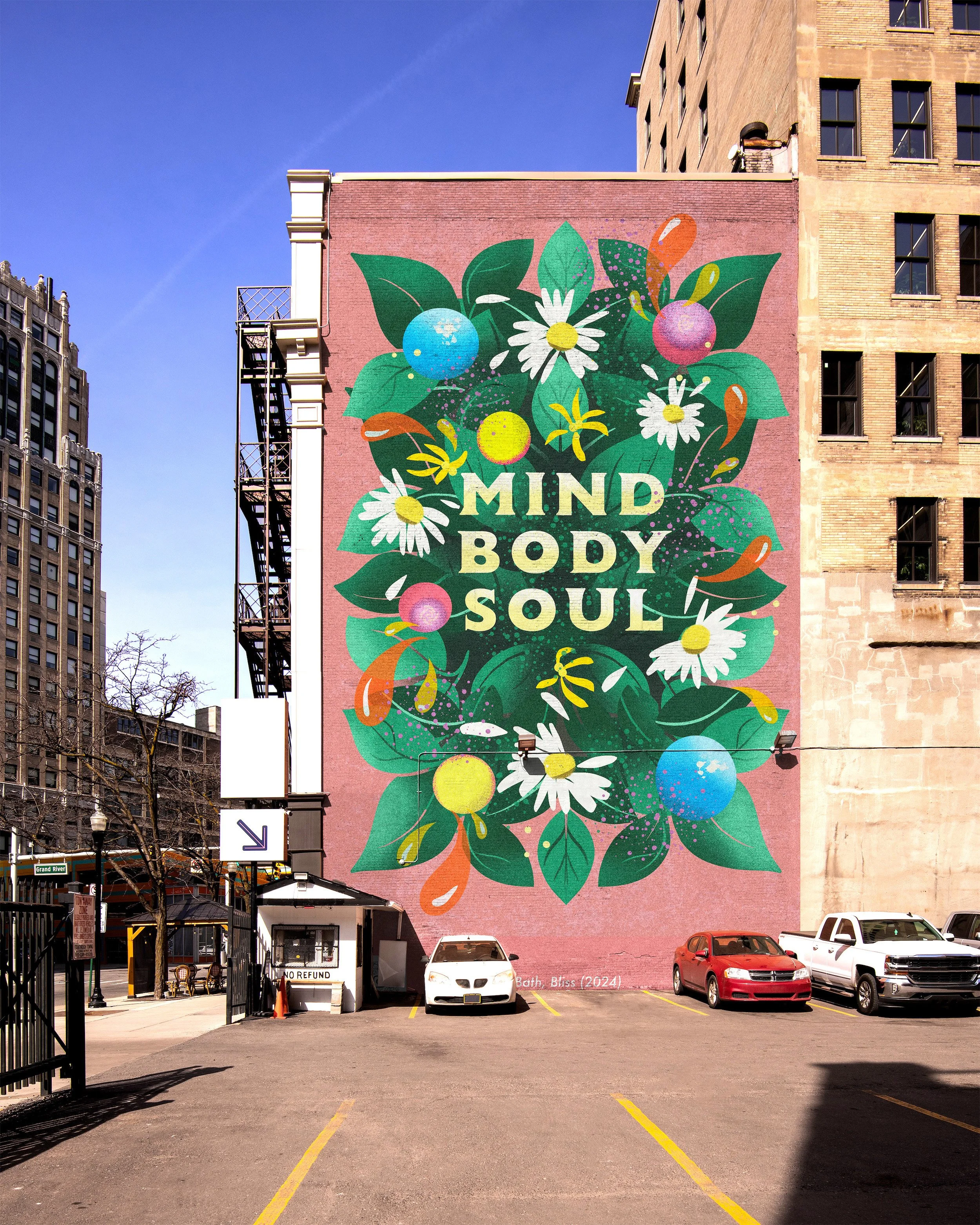 A large mural on the side of a building with a pink background, decorated with colorful flowers, leaves, and abstract shapes. The mural features the words "MIND BODY SOUL" in bold white letters. In front, there is a parking lot with several cars and a small booth with a sign that says "NO REFUND." Surrounding buildings are visible under a clear blue sky.