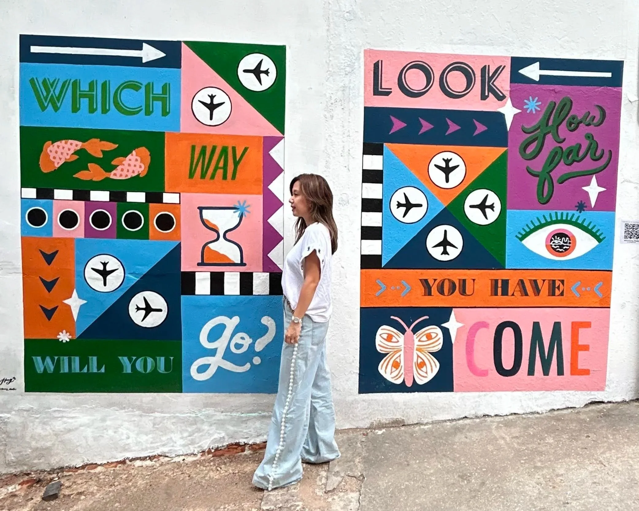 Colorful murals with text asking questions and making statements about directions, choices, and arrival, featuring symbols like airplanes, butterflies, an eye, and an hourglass. A woman standing in front of the murals.