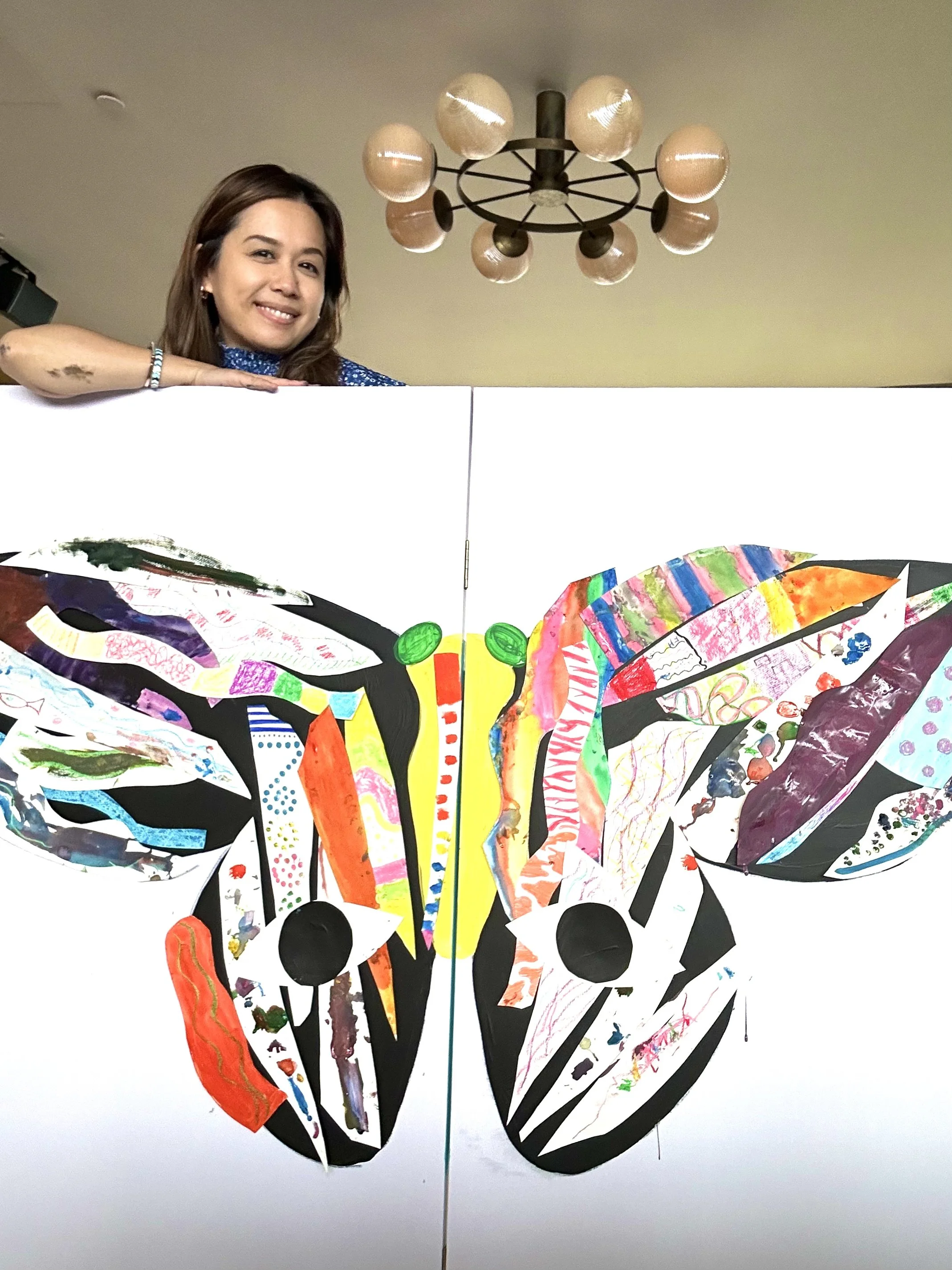 A woman smiling behind a butterfly artwork made from colorful paper and paint, with a round ceiling light fixture above.