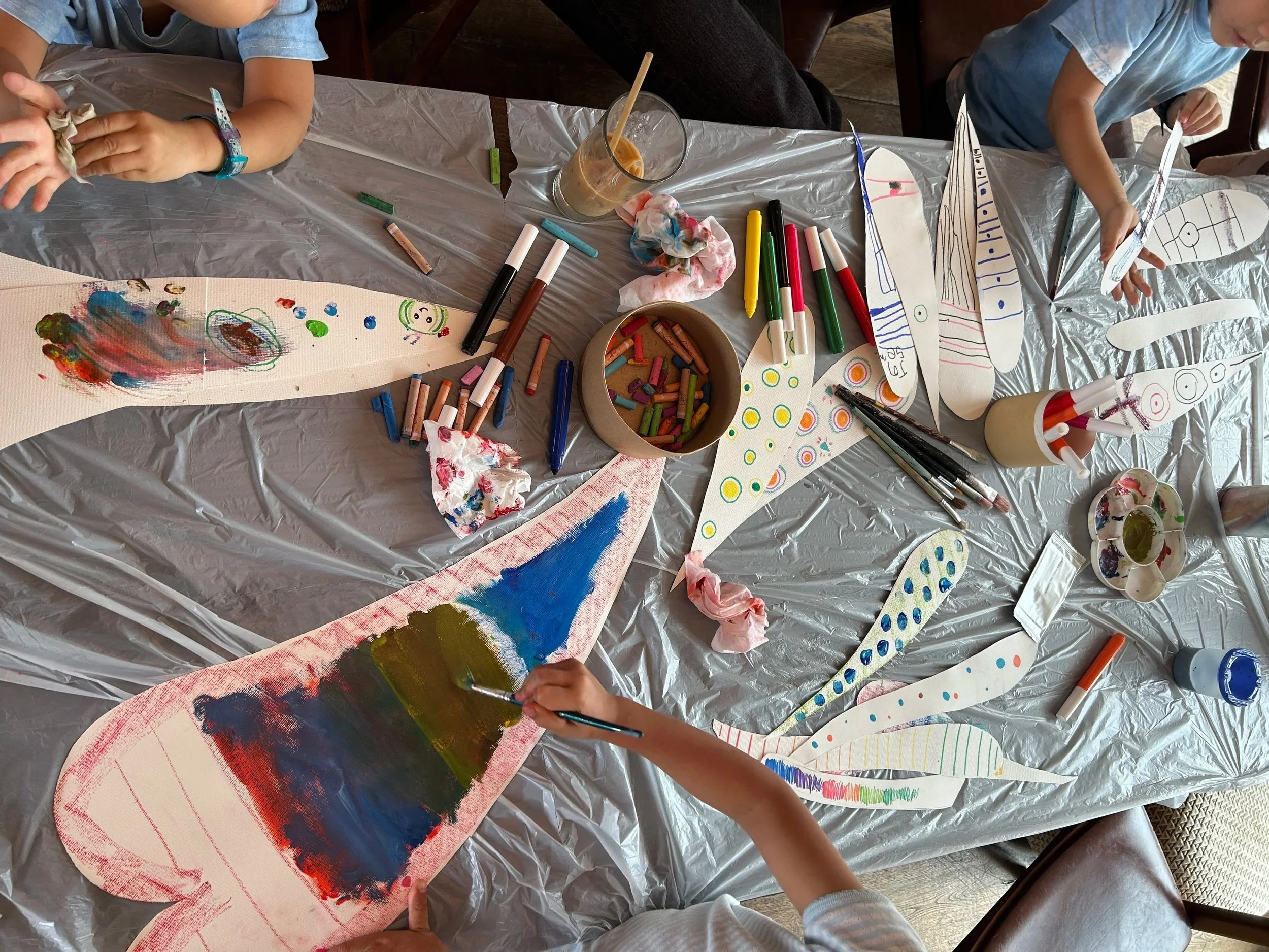 Children's hands painting and decorating paper dragonfly cutouts with markers and paints on a table covered with a plastic tablecloth, in Soho House