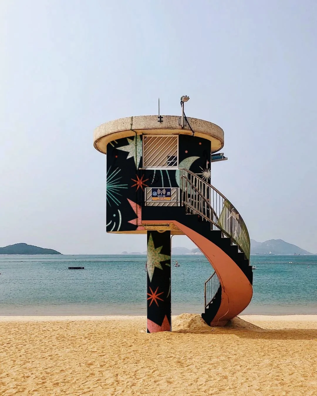 A lifeguard tower on a beach with a spiral staircase, decorated with colorful fireworks-themed artwork, overlooking the water with small islands in the background.