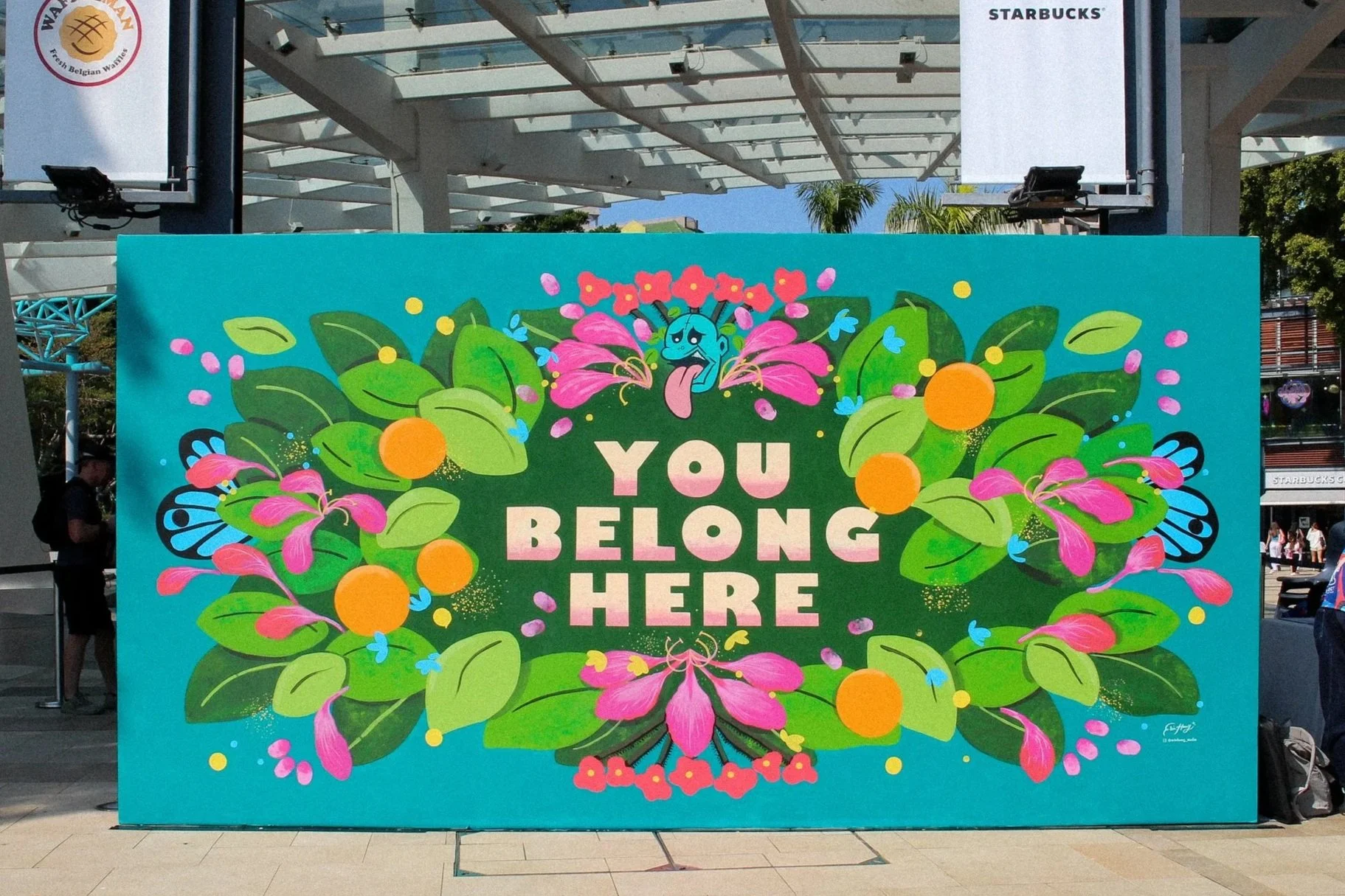 A colorful outdoor sign with floral and butterfly illustrations and the message "You Belong Here" in the center.