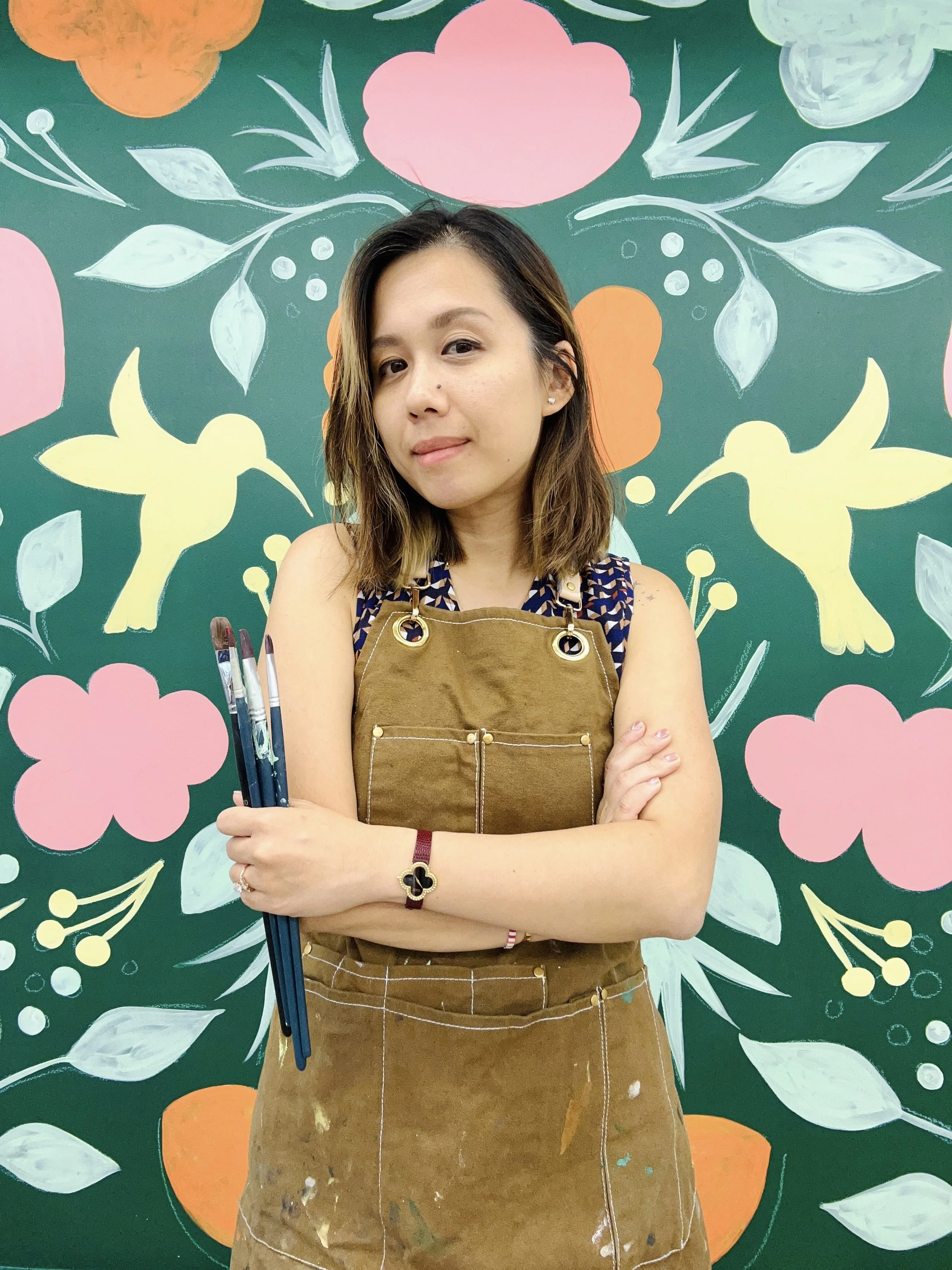 A woman with shoulder-length hair, wearing a painter's apron and holding paintbrushes, standing in front of a colorful floral and bird mural.