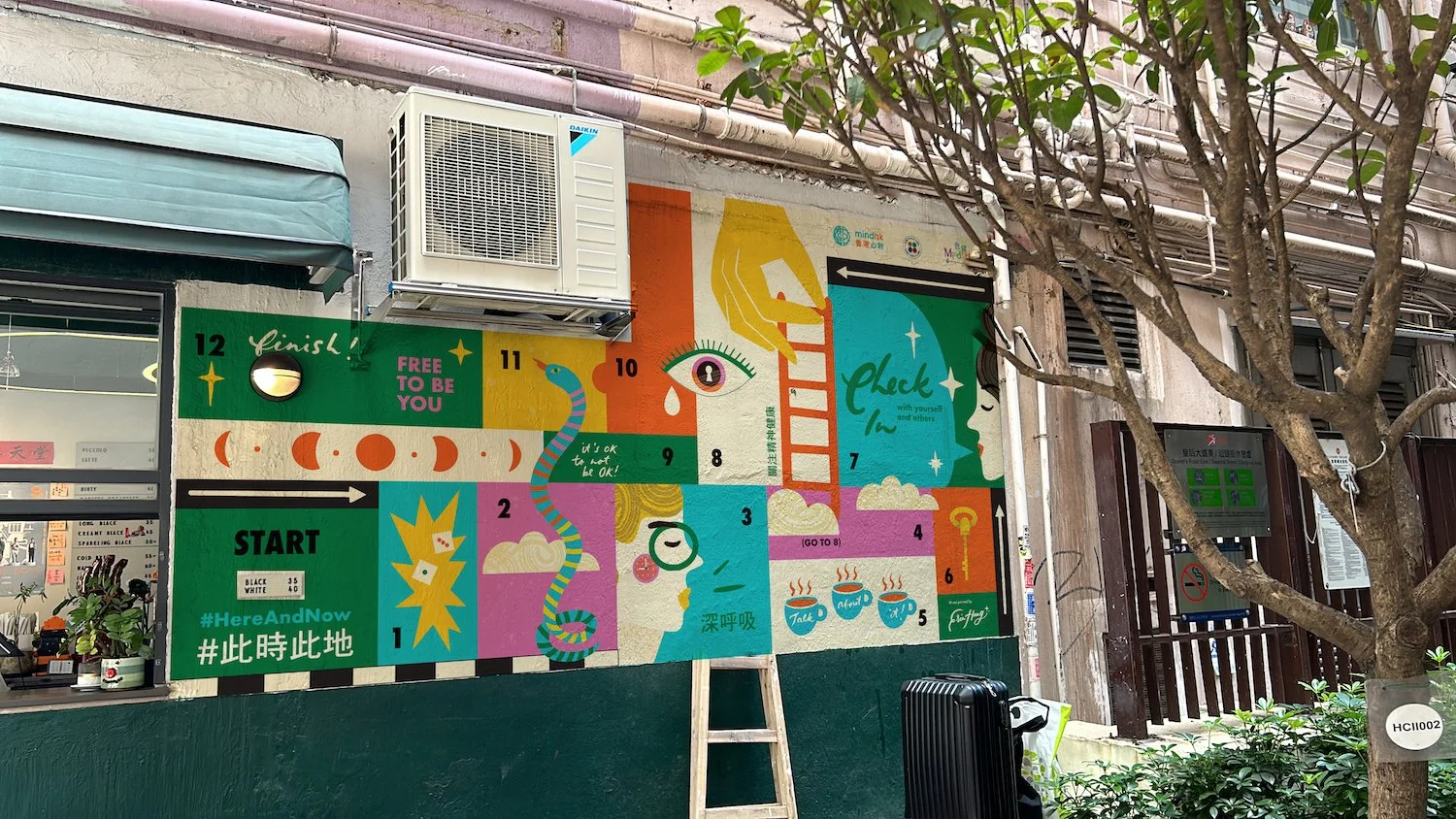 Colorful mural on a wall with a numbered game board, a snake illustration, a ladder, trees, and various icons including an eye, coffee cups, and a hand reaching for a ladder. The mural features vibrant colors and motivational phrases.