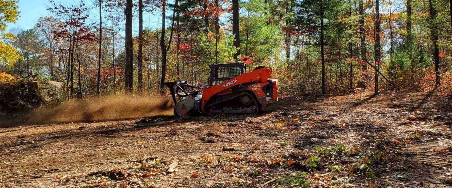 A red Kubota excavator with a snow blower attachment clearing dirt and leaves in a wooded area during daytime with fall foliage.
