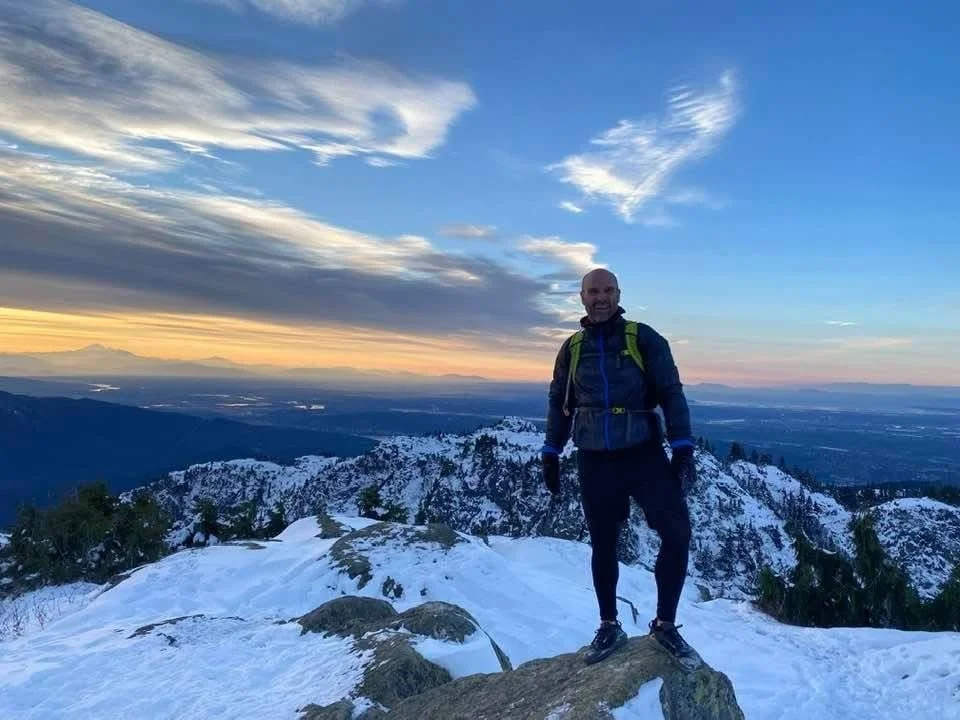 A man stands on snow-covered mountain rocks with a scenic view of a valley and mountains at sunset or sunrise, clouds in the sky, and a person with a backpack.