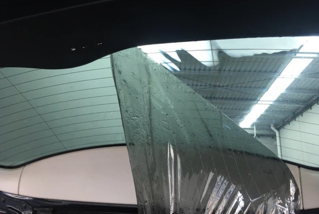 The image shows a car window with a large, triangular safety film or tint applied to it. The film is partially peeled away, revealing water droplets underneath and the reflection of the ceiling of a garage or workshop.
